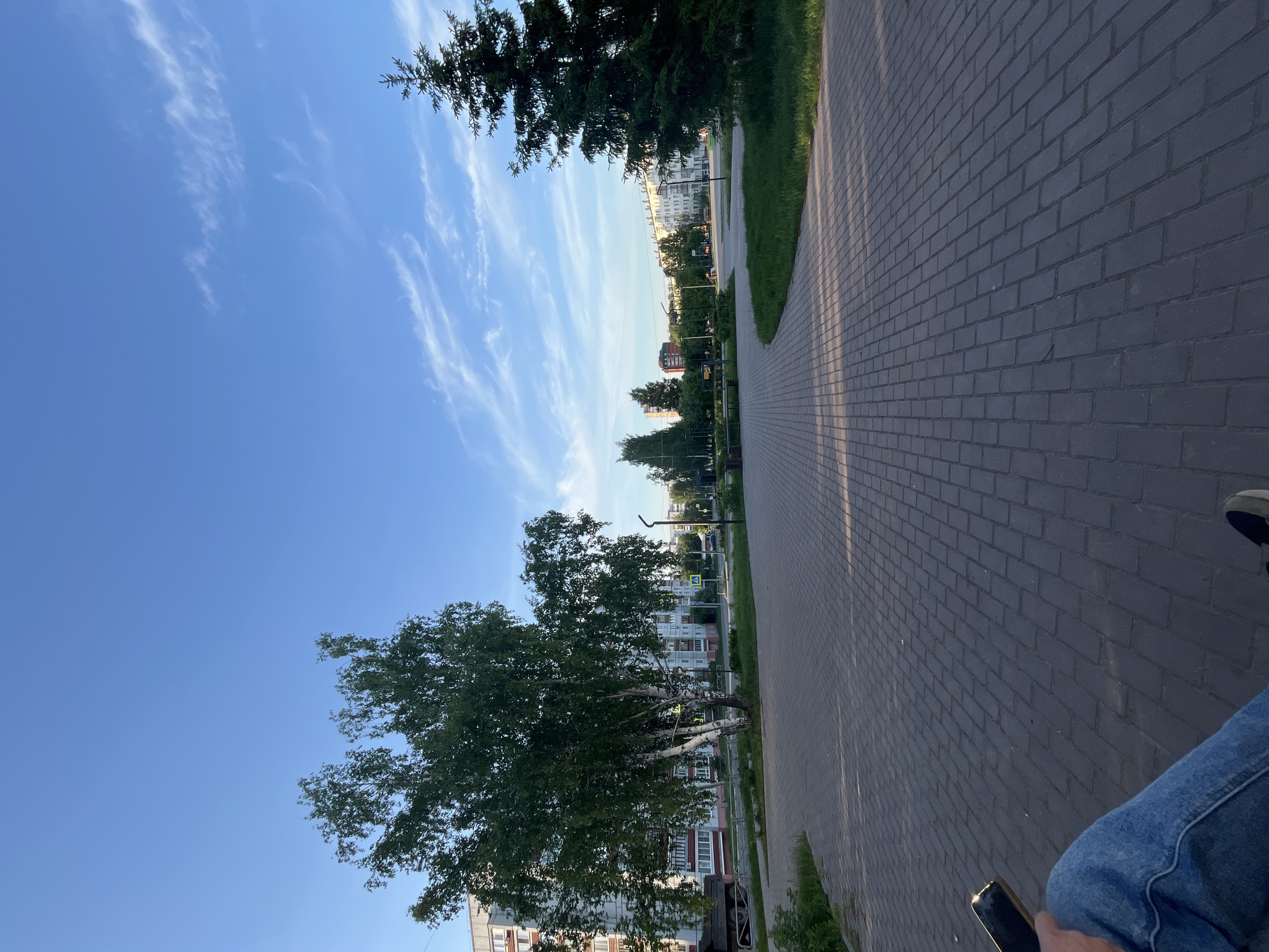 The image presents a view from a person’s perspective seated on a bench beside a brick walkway within a city park environment. The walkway stretches into the distance, lined with trees on both sides, and appears to be located in a residential area.
Environment:
A clear blue sky with wisps of cirrus clouds is visible. The lighting suggests either early morning or late afternoon. The ground is covered with grey brick pavers arranged in a standard running bond pattern. Patches of grass line the edge of the walkway on both sides. Buildings, mostly residential, can be seen in the background, indicating an urban park setting.
Objects:
A person’s leg, clad in blue jeans, is visible in the lower right corner. A mobile phone is being held by a hand with a small portion of one shoe visible. The walkway, trees, and buildings constitute the primary elements of the setting. A street light is also visible in the distance along the walkway.
Actions and Interactions:
The person holding the phone is seated. The perspective suggests observation.
Location:
Although the image does not explicitly state the location, the prompt references the city of Chaikovsky, Russia. Therefore, it is assumed that this park is located within this city.