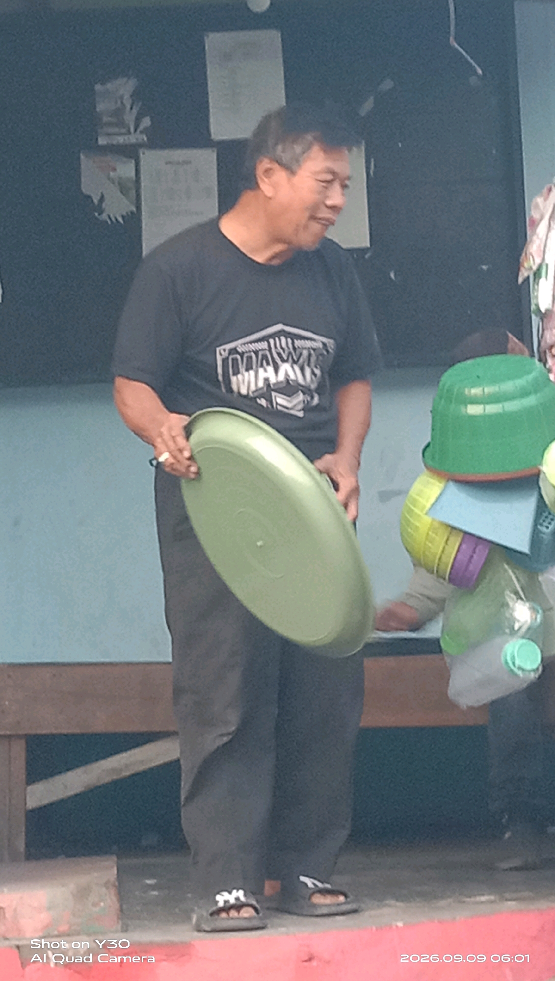 In Klangonan, Indonesia, a man wearing a black t-shirt with a "MAXIS" logo and dark pants is standing. He is holding a large, light green plastic basin. To his right, a stack of colorful plastic containers, including green and yellow basins and purple containers, is visible. The scene takes place outdoors, in front of a building with a dark facade. The ground appears to be paved with a reddish-orange surface. A wooden bench or railing is present in the background. The time stamp on the image indicates it was taken on September 9, 2026, at 06:01.