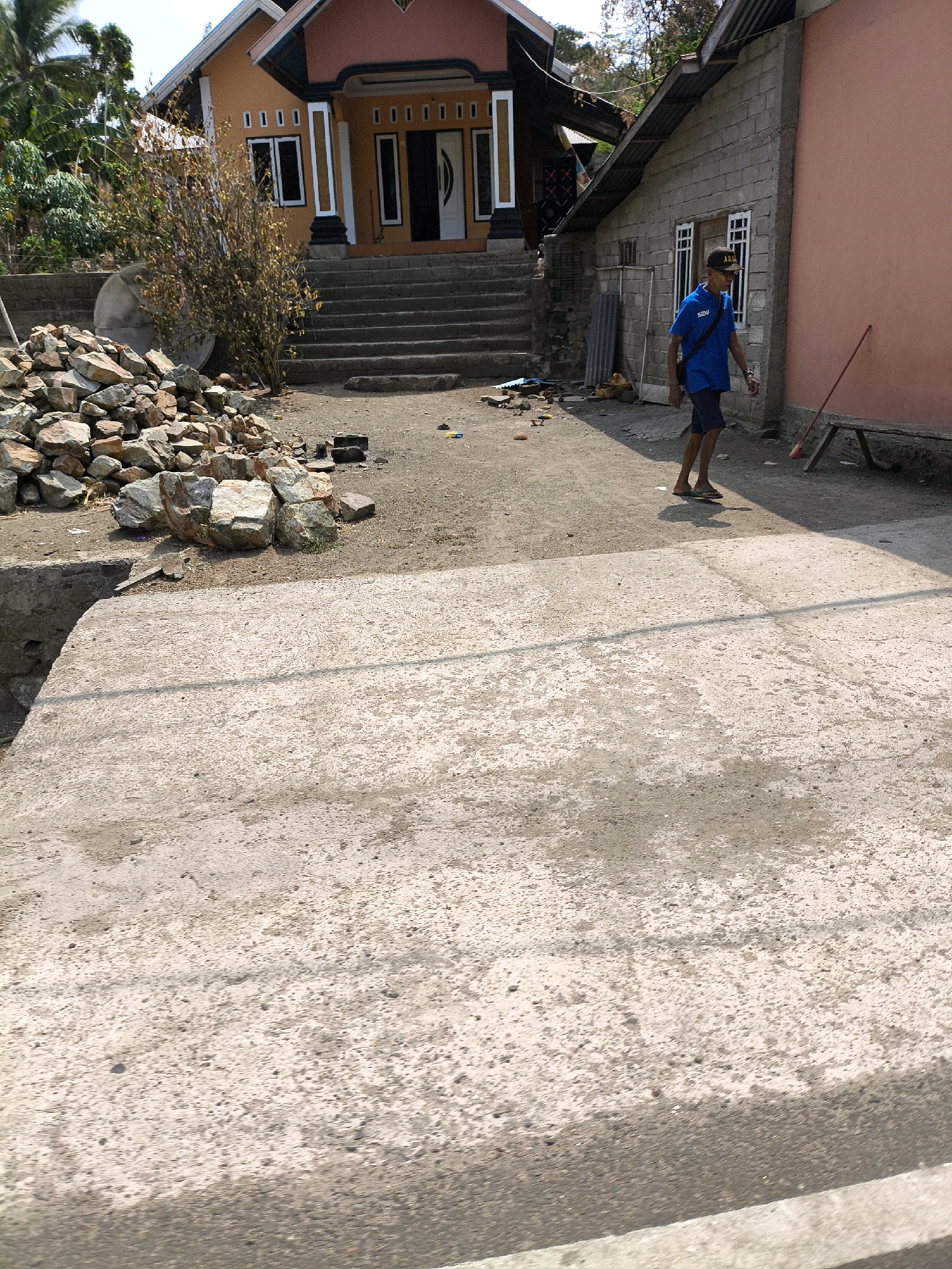 The image depicts a street scene in a residential area of an unknown city in Indonesia. In the foreground, a concrete walkway or road stretches into the distance. To the left, a pile of rocks and construction debris is visible. Behind the rocks, a house with an orange facade and a dark roof stands with a set of stairs leading up to the entrance. Adjacent to this house, a smaller, simpler building constructed with cinder blocks and a corrugated metal roof is present. A man wearing a blue t-shirt, dark shorts, a cap, and sandals is walking away from the viewer towards the right side of the image. He has a dark bag slung across his chest. A red broom rests against a wooden bench in front of the cinder block building. The environment appears somewhat unkempt, with loose dirt and scattered debris on the ground. The lighting suggests it is a sunny day.