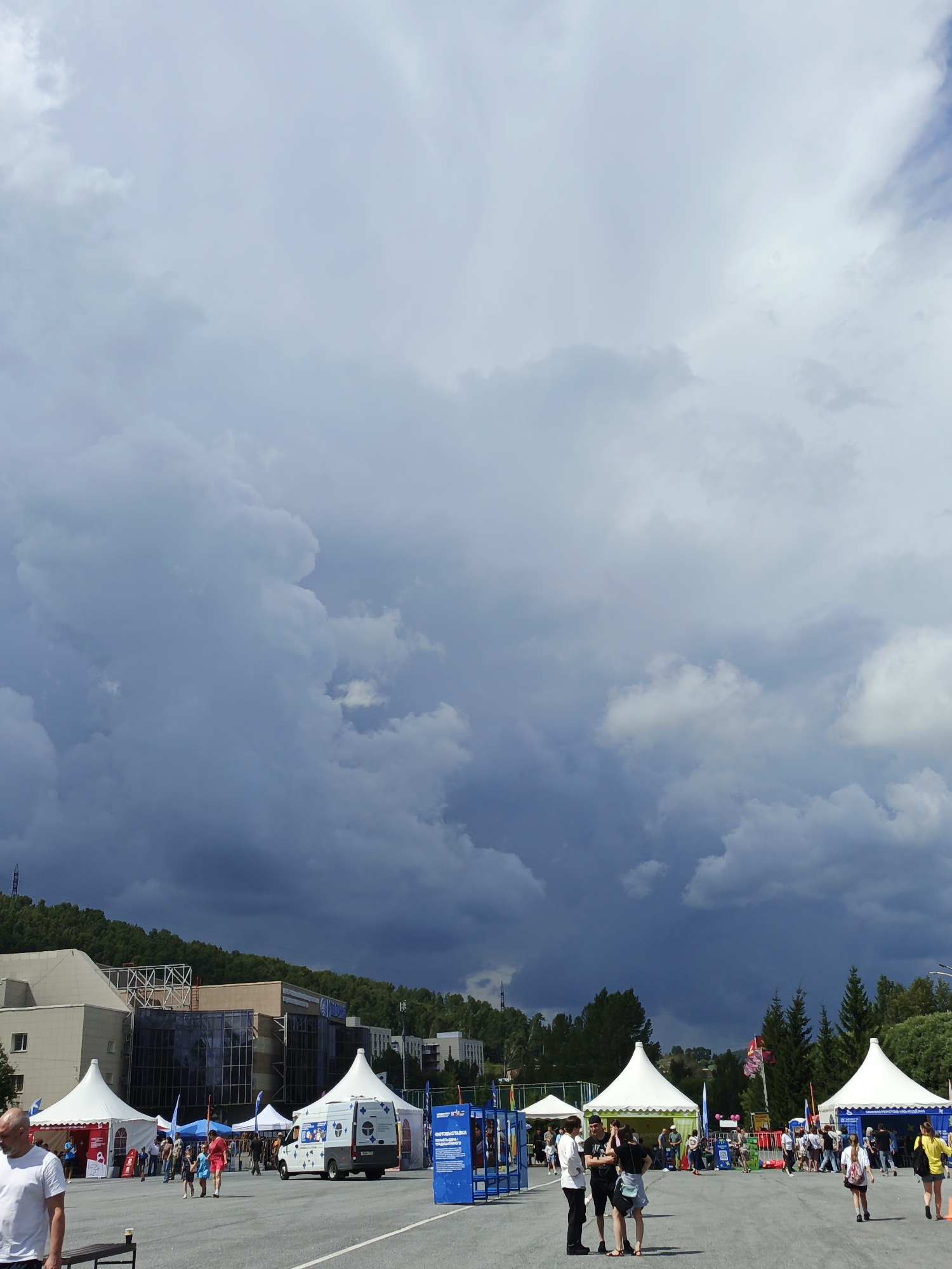 In Zlatoust, Russia, on a paved open area, several white tents are set up, suggesting an outdoor event or market. A white van is parked near one of the tents, with people visible milling around and interacting. In the background, there are buildings and a densely forested hillside under a dramatic, cloudy sky. The overall atmosphere is overcast, with dark storm clouds dominating the upper portion of the frame.