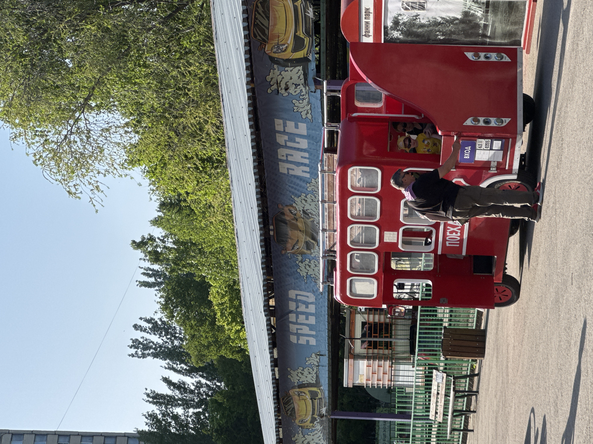The photograph shows a man interacting with a red double-decker bus-themed ride in an amusement park. The ride is decorated with the words "SPEED RACE" and images of cars, located next to a building or structure with a metal roof. The man, dressed in dark clothing, is standing near the entrance of the ride, appearing to be either assisting or observing children inside. The location is in a park, with trees and a partially visible building in the background. There are park benches nearby, and the ground appears to be paved. The image is taken in the city of Unknown, Unknown.