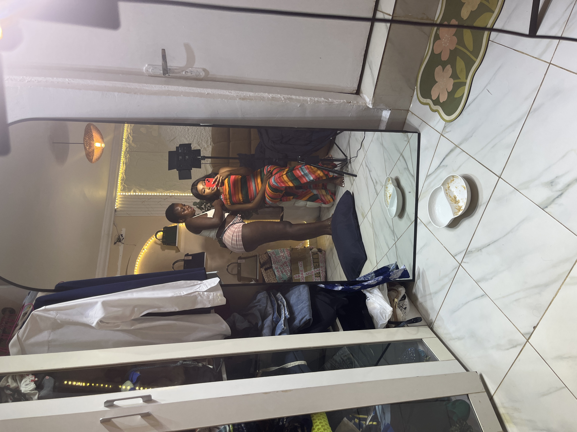 In a room with tiled flooring, two young Black women are visible in a full-length mirror. The woman on the left, wearing a white tank top and pink and white striped shorts, is standing and appears to be attending to the hair of the woman on the right. The woman on the right is seated and wearing a colorful striped maxi dress. She is holding a red smartphone, taking a selfie. In the background, a ring light is set up on a tripod. To the left of the mirror is a white wardrobe with open doors revealing clothes and storage. The overall environment suggests a personal space, possibly a bedroom or dressing area, located in Abuja, Nigeria.