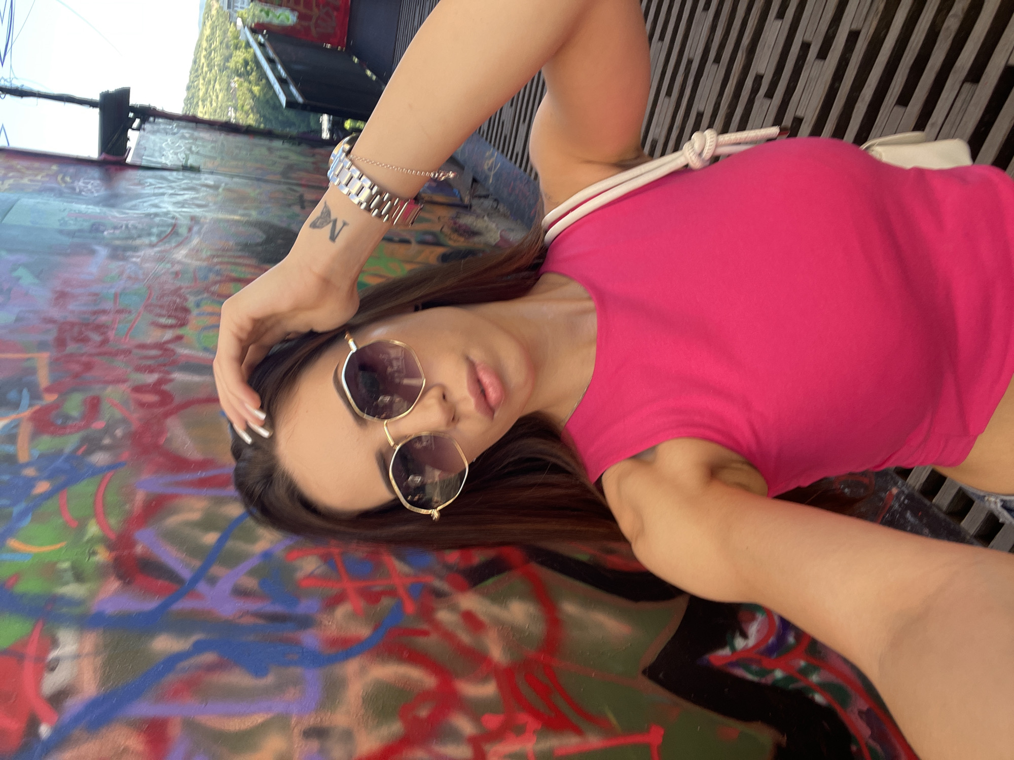 In the city of Unknown, Unknown, a young woman with long brown hair and a bright pink top is taking a selfie. She is wearing large, hexagonal sunglasses with gold frames and a silver watch on her left wrist, which also has a small butterfly tattoo. Her right arm is raised, with her hand resting on top of her head. The background is a vibrant graffiti-covered wall, with abstract splashes of color like red, blue, and purple. To the right of the woman, a metal railing and a grassy hillside with trees can be seen, suggesting an outdoor urban setting with a scenic overlook. A white crossbody bag with a knotted strap is visible on her right side. The ground beneath her appears to be a walkway made of dark, horizontal planks.