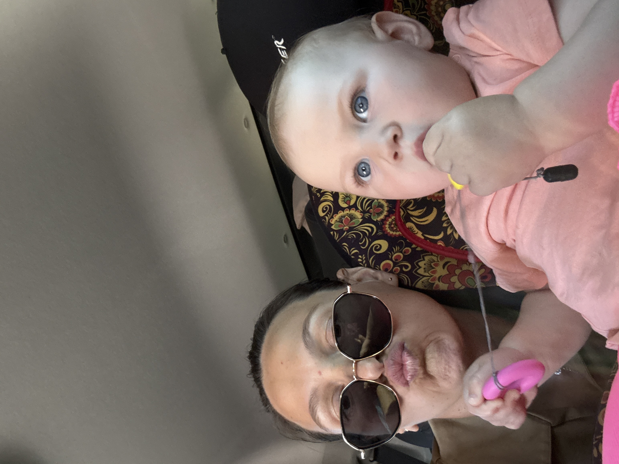 A selfie shows a woman with dark hair and sunglasses, and a baby with blue eyes. The baby wears a light pink shirt and holds a toy. They are positioned close together, likely in a car. The background is a light gray, possibly the interior of the vehicle. The scene appears to be indoors. There is no location information visible in the image.