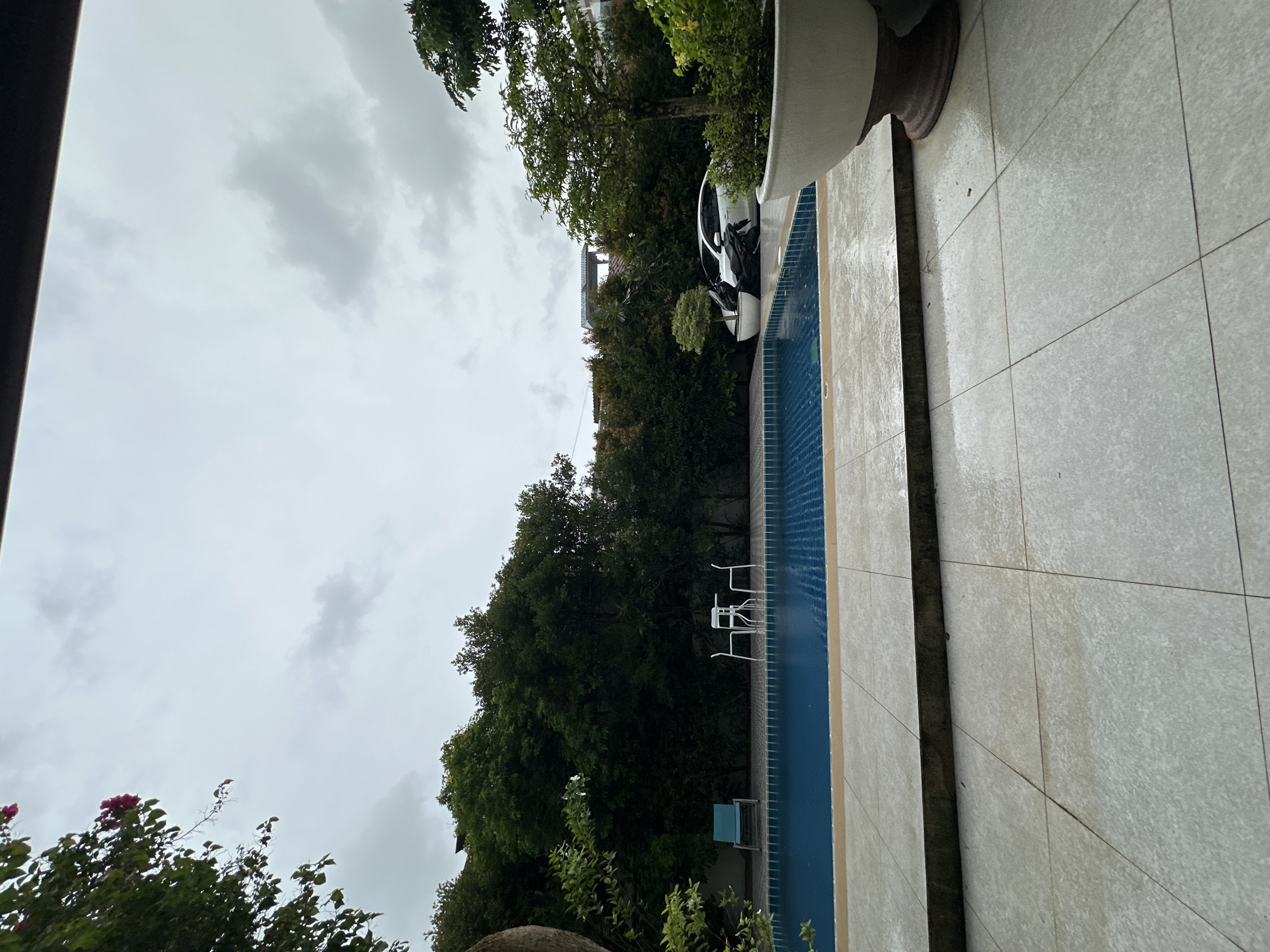 The image shows an outdoor swimming pool area on a cloudy day. The pool is rectangular with blue water and a tiled edge. To the left of the pool, there is a blue lounge chair and a small white table with two white chairs. Behind the pool, a dense wall of green trees forms the backdrop. To the right of the pool, there is a large white planter with a tree and other foliage. Further to the right, a white car and a motorcycle are parked. The foreground consists of a tiled patio area. The sky is overcast with gray clouds. The location is described as Unknown, Unknown.