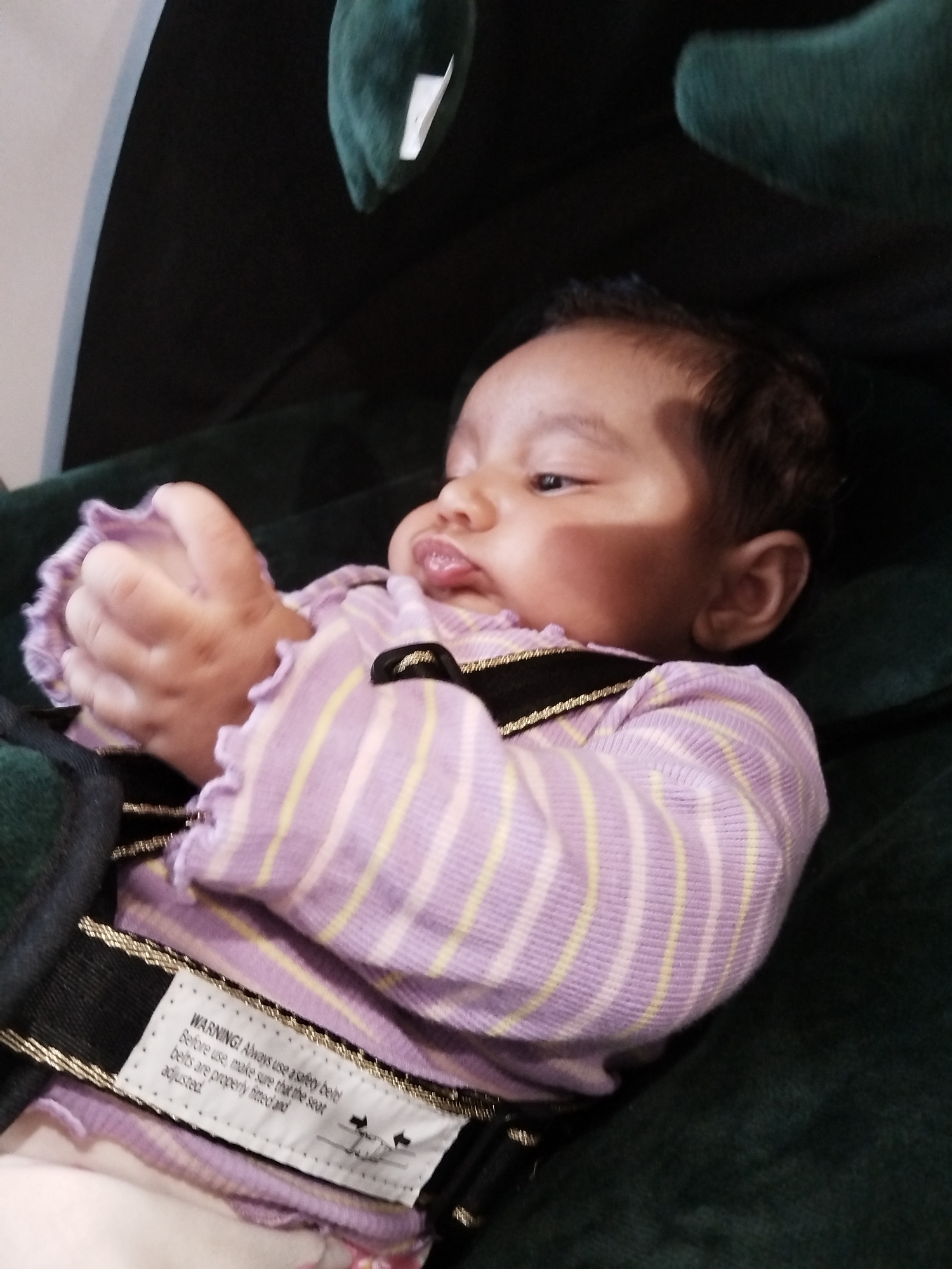 A baby wearing a light purple shirt with yellow stripes and secured with a black safety harness is lying on a dark green surface. The baby has dark hair and appears to be looking to the side with a slightly puckered mouth. Part of a label attached to the harness is visible, reading "WARNING! Always use a safety belt! Before use, make sure that the seat belts are properly fitted and adjusted." The location is Unknown, Unknown.