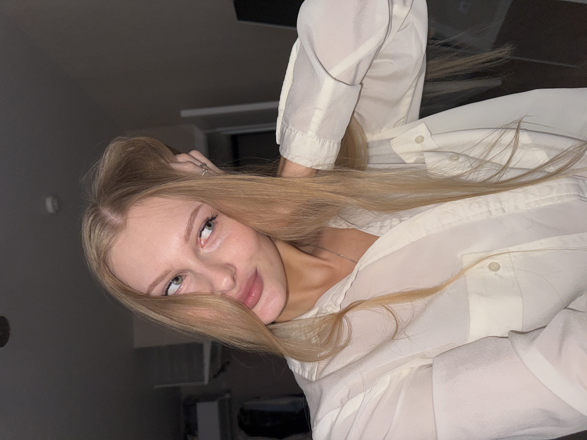 A blonde woman with green eyes and pale skin is shown in a close-up shot. She is wearing a white collared shirt with buttons and pockets. Her right hand is resting on her head, and her hair is blonde and long. She is looking to her left with a slight smirk. The background is dark and out of focus, with hints of furniture and what appears to be a closet. The image quality is good, with clear details of the woman's face and clothing. There is no indication of the specific location within the city of Perm, Russia.