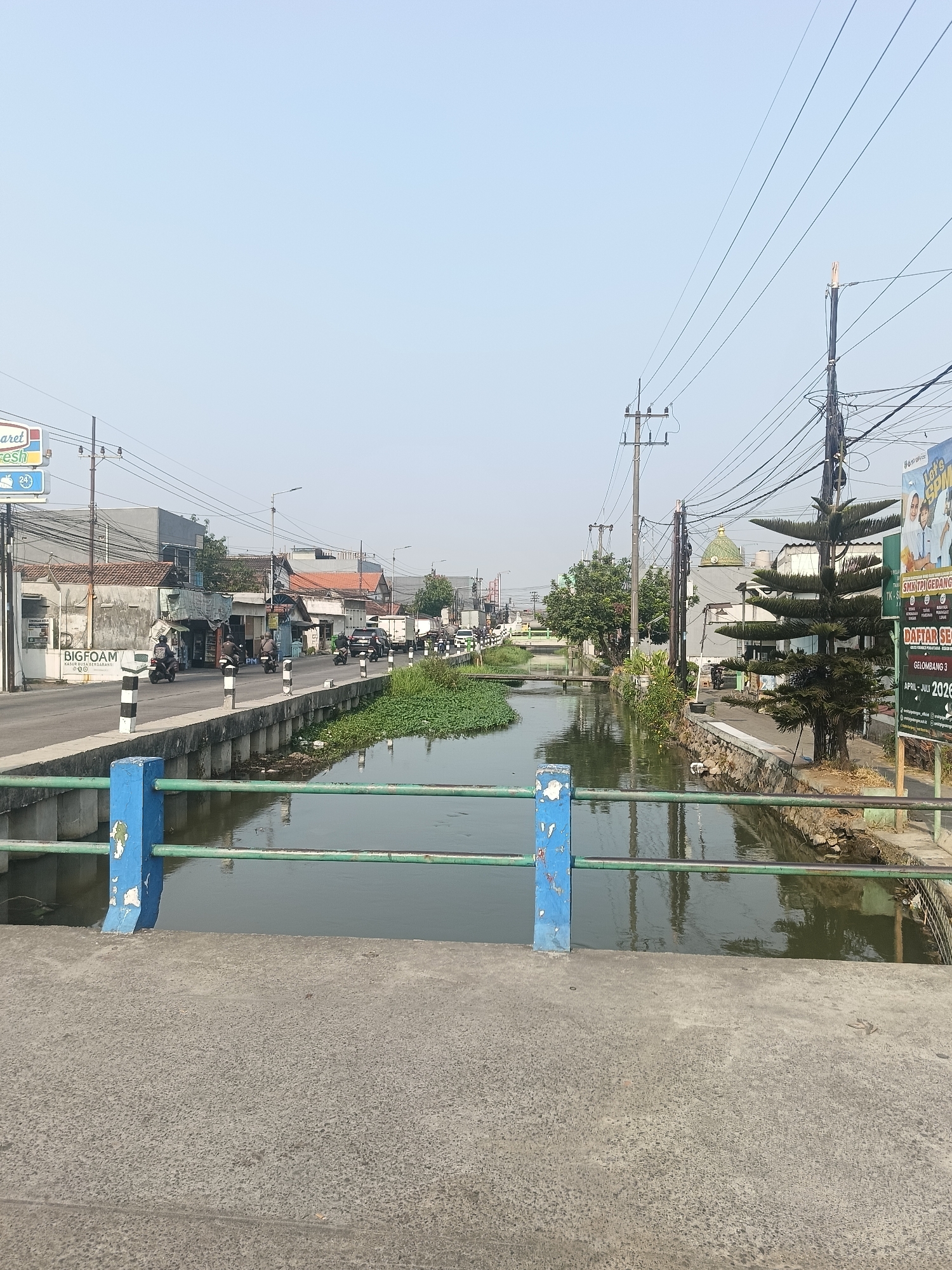 This is a photograph taken in the city of Wedi, Indonesia, likely on a sunny day, as indicated by the bright sky and clear lighting. The image captures a view of a canal with murky water, bordered by a concrete walkway on both sides. A green metal railing with blue supports runs along the foreground walkway, suggesting the photographer is standing on a bridge or elevated platform.
On the left side of the canal, a road is visible with several motorcycles being ridden by people in casual attire. Shops and buildings line the road, with signs indicating commercial activity. One prominent sign reads "BIGFOAM". Further down the road, traffic, including trucks and cars, is observed.
On the right side of the canal, a sidewalk is present, with a tall, dark green coniferous tree standing prominently. A billboard with an image of people and text in Indonesian is also visible. The environment appears to be a mix of urban and natural elements, with the canal running through a developed area. The overall impression is that of a typical street scene in a town or city in Indonesia.