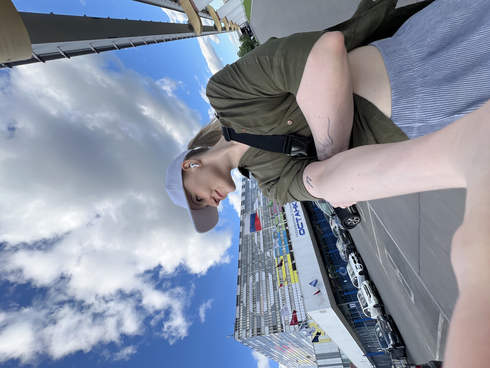 A medium shot shows a person standing in Khimki, Russia, with the Ostankino Technical Center tower in the background. The subject wears a white cap, a green button-down shirt, and light blue-striped shorts. A black strap crosses over their torso. Tattoos are visible on their arms. The sky is blue with scattered white clouds. The Ostankino Technical Center building is adorned with flags, and a row of parked cars lines the street next to it.
