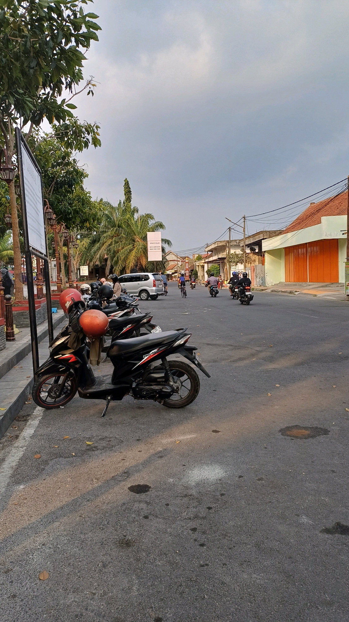 The image depicts a street scene in Klaten, Indonesia, likely during late afternoon or early evening given the long shadows cast across the asphalt road. In the foreground, several parked motorcycles, predominantly black scooters, are visible along the side of the road. Two of these motorcycles are equipped with orange helmets.
The road itself is paved and appears to be a two-lane street with some wear and tear, indicated by oil stains and a white line marking. Beyond the parked motorcycles, traffic is moving. A silver minivan is in the distance, and several individuals are riding motorcycles, some with passengers, further down the street. One person is seen riding a bicycle.
The environment suggests an urban or semi-urban setting. Buildings with tiled roofs line the right side of the street, featuring closed orange shutters on one structure. Palm trees and other leafy trees are interspersed with the buildings, contributing to a tropical feel. Overhead, a network of power lines is visible against a cloudy sky. A white banner, possibly an advertisement or information sign, hangs between two poles on the right side of the street. The overall atmosphere is one of everyday life and activity on a street in Indonesia.