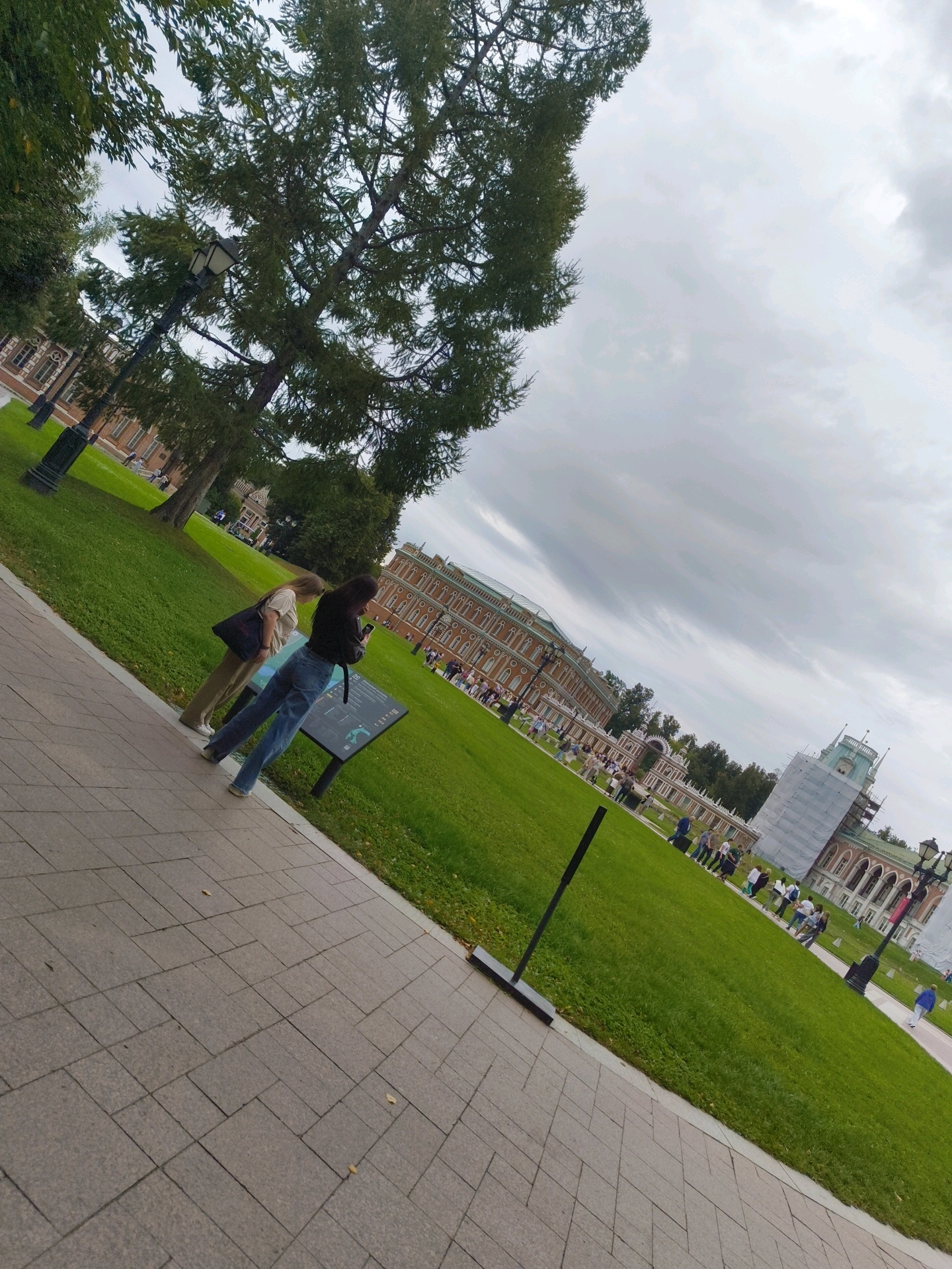 The image depicts an outdoor scene at the Tsaritsyno Museum-Reserve in Moscow, Russia. A paved walkway occupies the foreground, leading to a large expanse of green grass. In the middle ground, two women are standing near an information display. One woman, with blonde hair, wears a light-colored top and dark pants, and carries a tote bag. The other woman, with dark hair, is dressed in a black top and blue jeans, and is looking at her phone. Beyond them, a long, ornate building with a red facade and white architectural details is visible, characteristic of Russian imperial architecture. Further in the background, more buildings and trees are present, with a cloudy sky overhead. Several other people are scattered throughout the park, walking and observing the surroundings. One building on the right side of the frame is partially covered in scaffolding, suggesting ongoing renovation or construction. The overall atmosphere appears to be that of a public park on an overcast day.