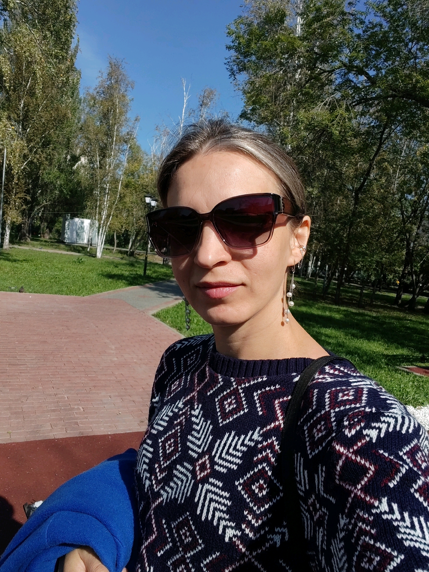 A woman, appearing to be in her late 20s to early 30s, is captured in a selfie taken outdoors. She is wearing large, dark-rimmed sunglasses that obscure her eyes, and a navy blue sweater with a white and burgundy geometric pattern. Long, delicate earrings with pearls hang from her ears. Her hair is light brown with blonde highlights, pulled back from her face. The background suggests a park setting with trees, green grass, and a paved walkway made of red bricks. The sky is clear and blue, indicating a sunny day. A bright blue folded garment, possibly a coat or scarf, is visible in the lower left of the frame, held by her hand. The overall lighting indicates it is daytime.