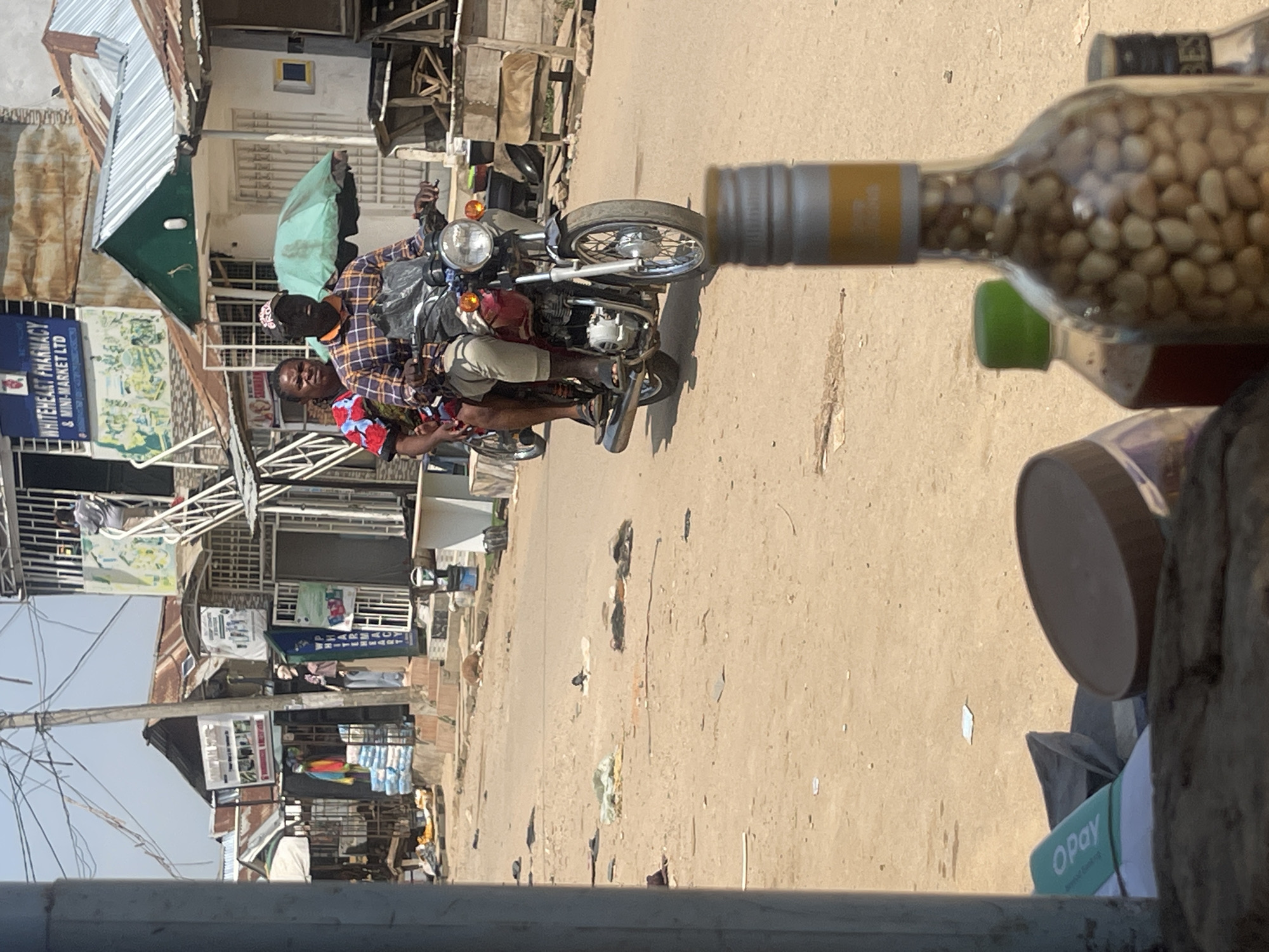 In Abuja, Nigeria, a motorcyclist wearing a plaid shirt and a cap drives a red motorcycle down a dusty street. A woman with a patterned top sits behind him. Several buildings with corrugated metal roofs line the street, some with signs for businesses such as "WHITEHEART PHARMACY & MINI-MARKET LTD." In the foreground, a blurred bottle filled with nuts sits on a surface. The street is unpaved and scattered with debris.