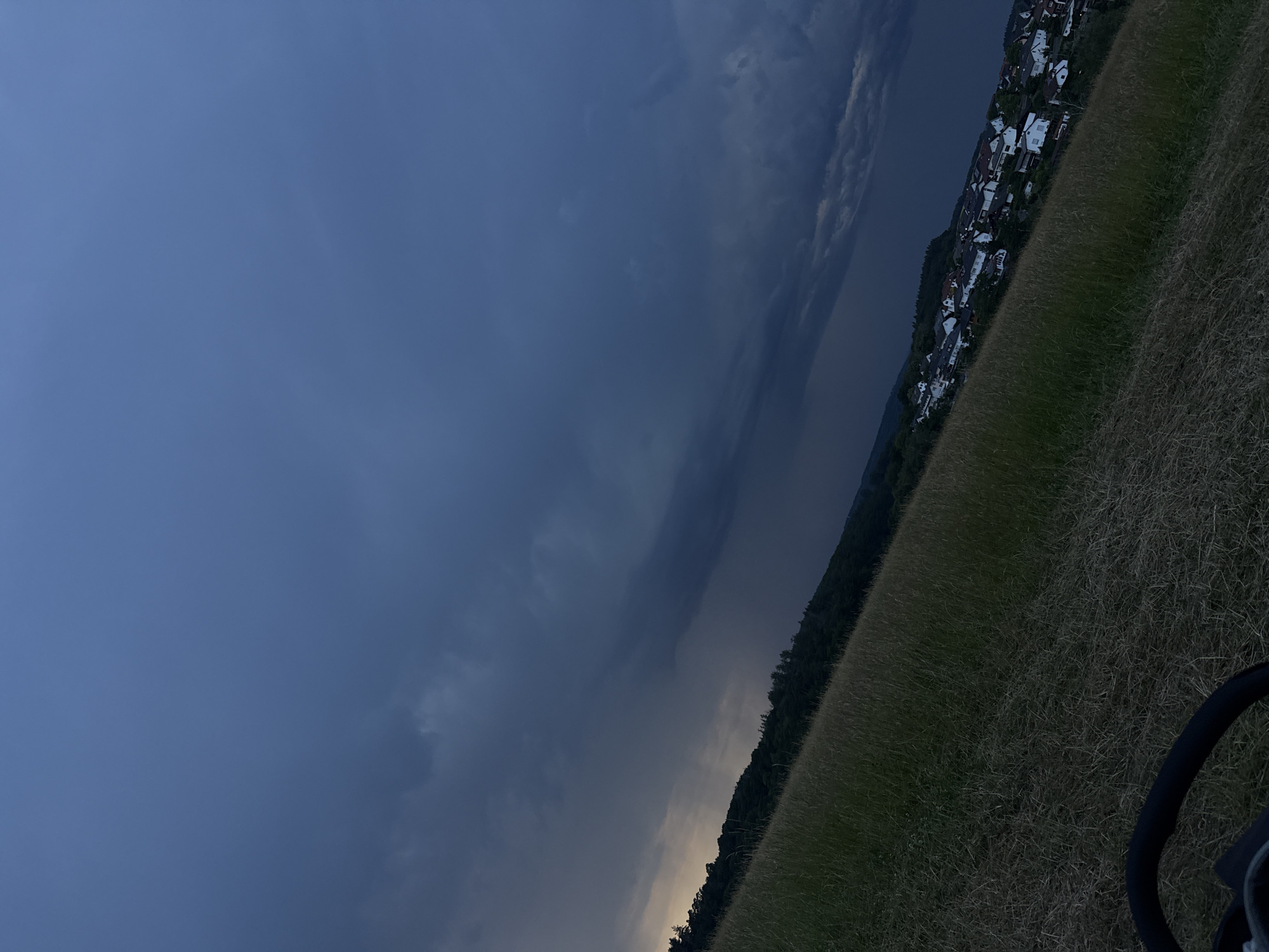 The image depicts a twilight scene in Niederquembach, Germany, with a dramatic, dark sky filled with storm clouds. The foreground is a sloping field of dry grass. In the midground, a dense line of trees separates the field from a village nestled on the hillside. The village consists of numerous residential buildings with white walls and dark roofs. The overall impression is of an impending storm over a rural landscape. There are no people or animals visible in the image. A portion of what appears to be a stroller or pram handle is visible in the bottom left corner of the frame.