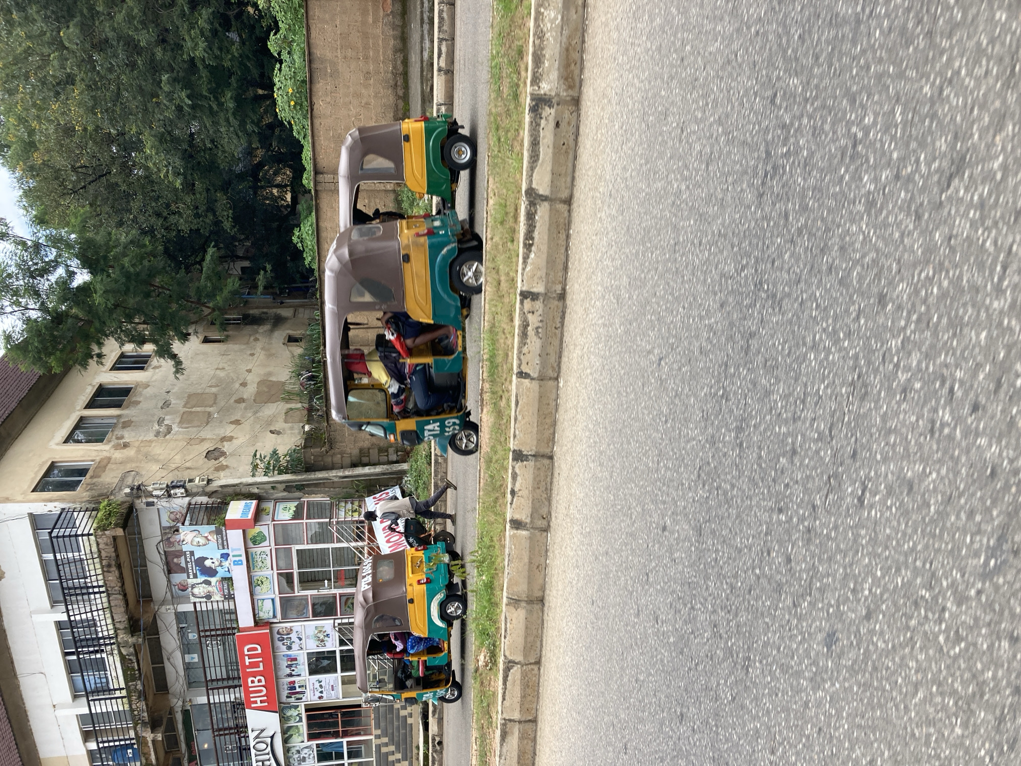 In Jos, Nigeria, a street scene unfolds with a focus on three motorized tricycles, commonly known as "Keke Napep." These vehicles are a common mode of transport in the city. The tricycles are colorful, predominantly yellow and green, with brown roofs. Two of the tricycles are positioned next to each other on the road, with the third slightly behind and to the right. The tricycles are carrying passengers, their faces partially obscured. One of the Keke Napep has the license plate "PTA-459". A man in a light-colored shirt and dark pants is walking on the road, crossing in front of the two closer tricycles.
The backdrop of the scene is a two-story building with a weathered facade, suggesting it's an older structure. The building has several windows and balconies, and some signage can be seen on its lower level. One sign reads "FASHION HUB LTD". Lush green trees and vegetation are visible behind the building, indicating a somewhat green environment despite the urban setting. The road surface appears to be asphalt, with a concrete curb and a grassy verge separating it from the sidewalk or area in front of the building. The overall impression is a slice of everyday life in Jos, highlighting the ubiquitous presence of Keke Napep and the blend of commercial and residential elements.