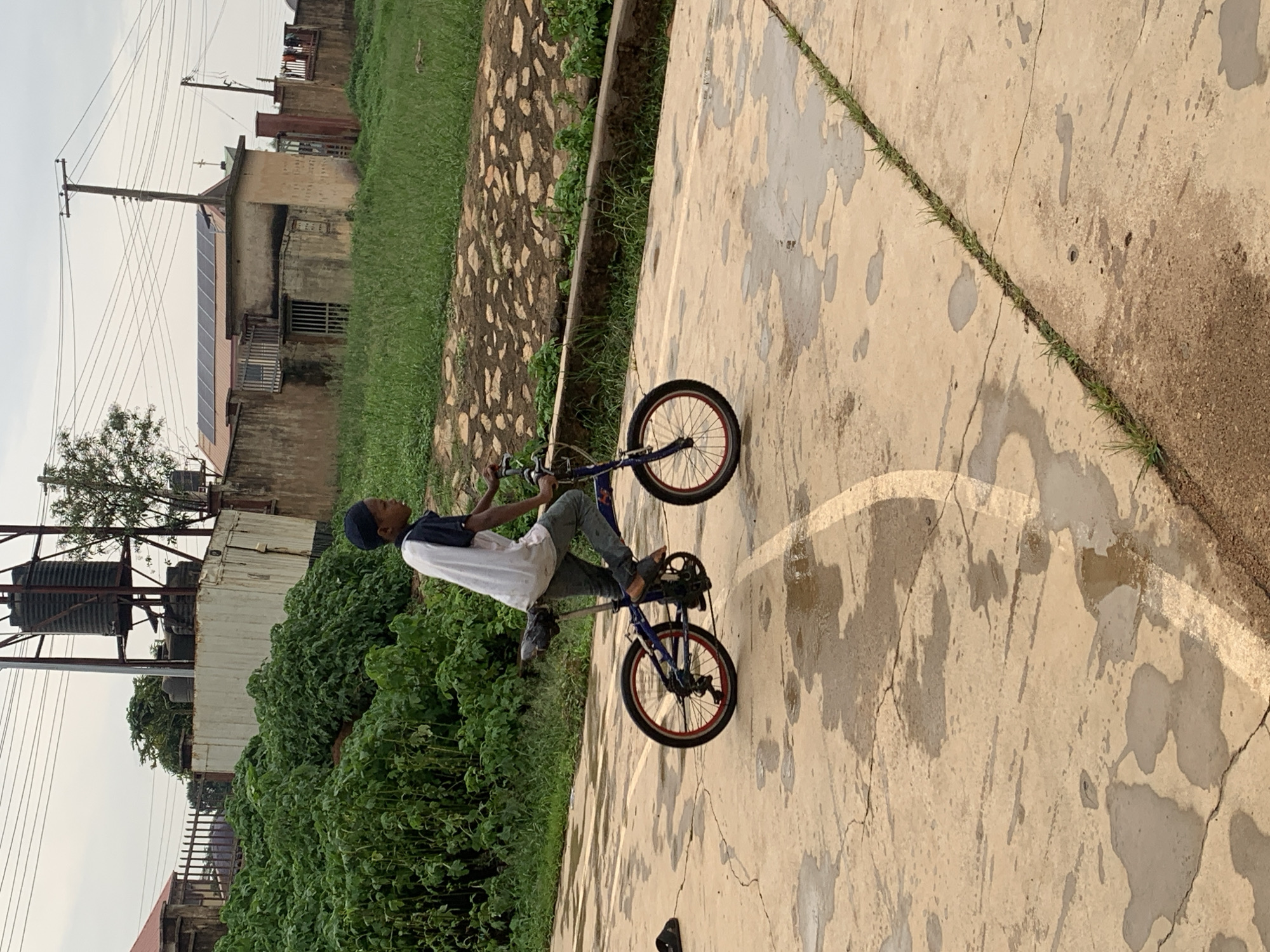 A young boy wearing a blue baseball cap, a white t-shirt with navy blue accents, and dark trousers rides a blue folding bicycle with red-rimmed wheels on a wet, concrete outdoor surface. The boy is in motion, with his feet on the pedals and hands on the handlebars. To the left of the boy, a black sandal lies on the ground. In the background, a dense green bush is visible, followed by a concrete wall and some buildings. A water tank is mounted on a metal structure behind the bush. Overhead, power lines crisscross the sky, and the weather appears overcast. The environment suggests an outdoor space in an urban or semi-urban setting, possibly a courtyard or a pathway in Gura Top, Nigeria.