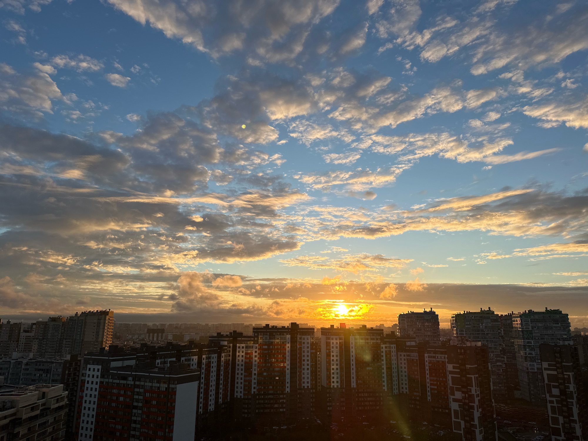 This is a high-angle, wide shot of a city skyline during sunset. The sky is filled with dramatic clouds illuminated by the setting sun, which appears as a bright, golden orb near the horizon. The clouds range in color from white and yellow to gray and blue, with shafts of sunlight breaking through in some areas. In the foreground, a dense collection of modern apartment buildings dominates the view. These buildings are characterized by their multi-story structures, varied architectural styles, and numerous windows. Some buildings are dark and silhouetted against the bright sky, while others catch the warm light of the sunset, revealing their colors and details. The cityscape extends into the distance, with more buildings faintly visible under the hazy sky. The overall impression is that of an urban environment at dusk, with the sky providing a vibrant and dynamic backdrop to the residential structures. The specific location of Novaya Pustosh, Russia, is not visually discernible from the image itself, as the visual elements are generic to many modern cityscapes.