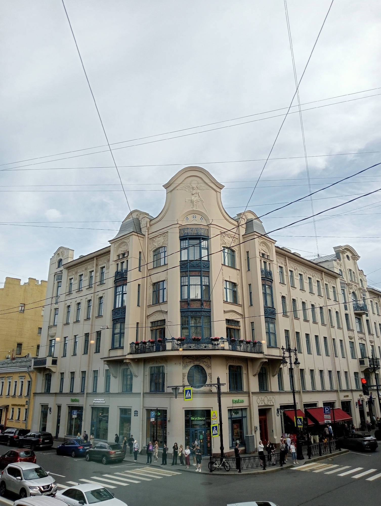 This media file shows a street scene in Saint Petersburg, Russia, on an overcast day. The foreground features a street with several cars, including a silver SUV and a white sedan. A pedestrian crossing is visible, with a few people standing on the sidewalk. In the background, a large, ornate building with cream-colored walls and blue-trimmed windows dominates the view. The building has multiple stories, balconies, and decorative architectural elements. A yellow building is partially visible to the left of the cream-colored building.
Several people are walking on the sidewalk in front of the building. There are also signs visible, including a red traffic light and parking signs. Text on the cream-colored building includes "ВкусВилл" (VkusVill) and what appears to be an address or business name in Russian. Overhead, a complex network of electrical wires crisscrosses the sky. The overall impression is a bustling urban environment under a cloudy sky.