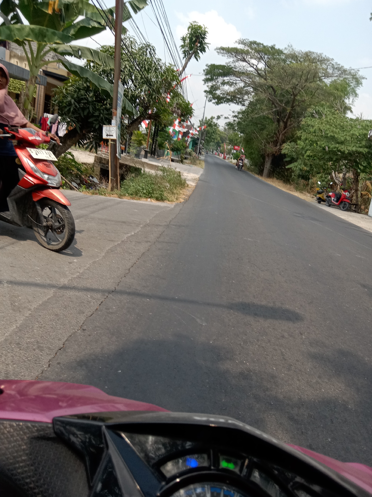 The image captures a street scene in Pacing, Indonesia. A person on a red scooter is visible on the left side of the road, partially obscuring the foreground. The scooter has a license plate that reads "S 2017 AA8". The road itself is paved and appears to be cracked in places. On the right side of the road, there are several trees and some vegetation. In the background, there are more trees, some houses, and power lines running overhead. Further down the road, two other scooters are parked on the side, one red and one yellow. The sky is clear with some clouds. The scene suggests a typical day in a semi-rural or suburban area of Indonesia.