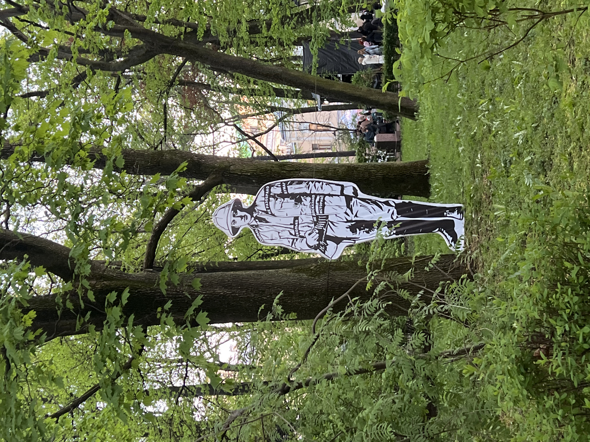 The image depicts a park in Saint Petersburg, Russia. A black-and-white cardboard cutout of a soldier stands amongst trees and green shrubbery. A small crowd of people is gathered in the background, possibly for an event. Buildings are visible behind the crowd. The setting appears to be outdoors on a bright day with lush foliage.