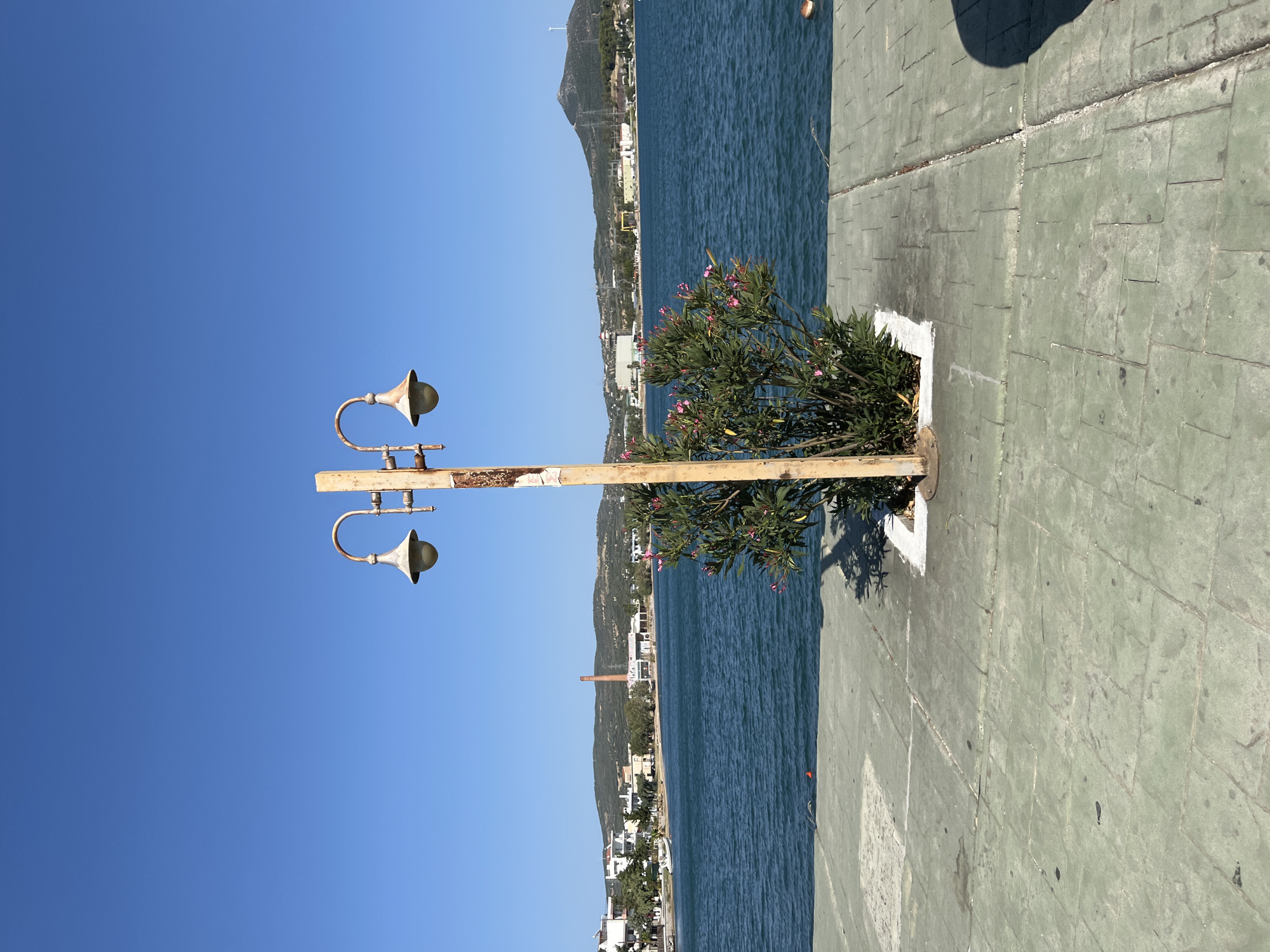 The image depicts a seaside scene in Karavos, Greece, under a clear blue sky. A weathered, single-post lamppost with two lamps stands in the foreground, centered in the frame. A small bush with pink flowers is planted at the base of the lamppost, enclosed by a white border. The ground in the immediate foreground is a paved area made of large, square concrete slabs with a greenish tint, showing some wear and tear. Beyond the paving, a body of calm, dark blue water stretches to the horizon.
Across the water, the coastline is visible, featuring a town with buildings in white and beige, nestled against rolling green hills. A prominent red brick smokestack rises from a building complex on the coast, suggesting an industrial presence. In the distance, the landscape is characterized by hills and mountains, with a wind turbine visible on the crest of one of the larger hills. The overall impression is one of a tranquil coastal environment with a touch of urban development along the shoreline.