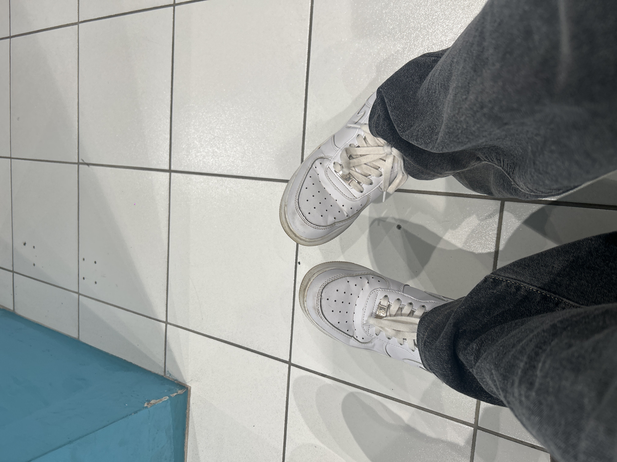 An overhead, slightly angled shot captures a person's feet and lower legs, adorned in white athletic shoes and dark gray jeans, standing on a white tiled floor. The shoes, resembling Nike Air Force 1s, feature perforated toe boxes and white laces, showing signs of wear and some dirt. The dark gray jeans are loose-fitting, creating folds around the ankles.
The floor is composed of large, light-colored square tiles with dark gray grout lines. Shadows are cast by the feet and legs, indicating an overhead light source. In the bottom left corner, a low, light blue structure, possibly a painted concrete block or bench, is visible.
The setting appears to be an indoor or covered public space, such as a mall, a large hall, or a public building, characterized by its utilitarian tiled floor and plain aesthetics. The lighting suggests it is daytime or a well-lit interior. There are no specific activities or emotions discernible. No visible text is present within the image that could offer further context. While the location context is provided as Saint Petersburg, Russia, the visual content of the image itself does not contain any specific cues to confirm this geographical location.