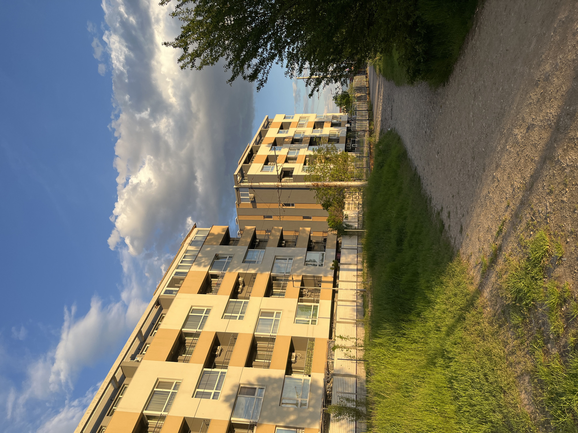 The image captures a view in Sofia, Bulgaria, showing two modern residential buildings under a partly cloudy sky. The buildings are characterized by their beige and gray facade, with multiple windows and balconies. A dirt path runs alongside the buildings, bordered by green grass. A tree is partially visible in the upper right corner. The perspective is slightly tilted, giving a dynamic angle to the scene. No people are visible in the frame.