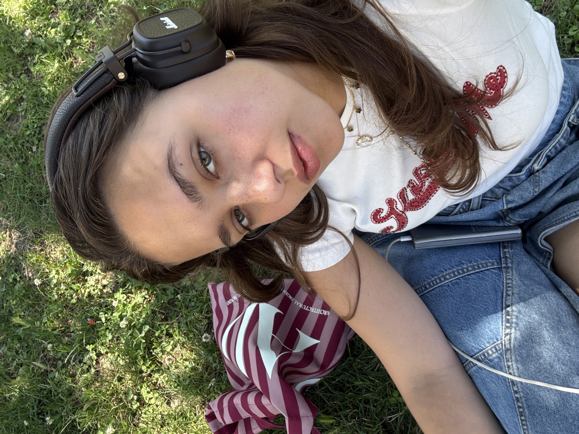 A young woman is lying on her back in a grassy area, wearing large black headphones and a white t-shirt with red sequined embellishments. She has brown hair and is looking upwards towards the camera. Her makeup includes lip gloss and mascara. She is wearing a gold necklace with a pendant. Her legs are crossed, and she is wearing denim shorts. A striped bag with the letters "AD" is visible next to her. A silver power bank is resting on her lap, connected by a white cable. The scene appears to be outdoors, with natural lighting suggesting it is daytime. The grass has small white flowers scattered throughout. The overall mood is relaxed and casual. Some text is visible on the striped bag: "ARCHITECTURAL DIGEST" and possibly "DOMINICO".