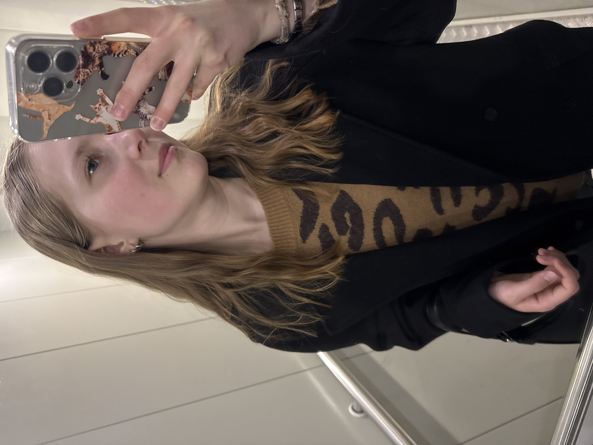 A young fair-skinned person with light brown hair is taking a selfie with an iPhone encased in a clear case featuring cat designs. The person wears multiple bracelets on their left wrist and a small hoop earring in their left ear. They are wearing a brown sweater with a leopard print design and a black coat. The background appears to be a room with white walls and a chrome handrail. The location is difficult to determine from the image and cannot be placed in Chaikovsky, Russia with the information provided.