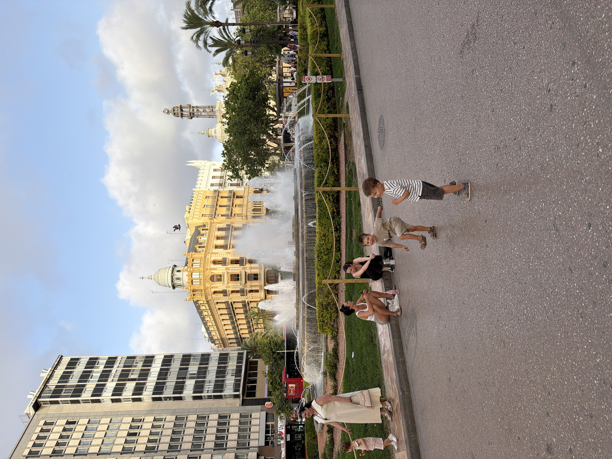 In an urban setting, a large fountain with multiple jets of water is the central focus. The fountain is surrounded by a manicured hedge and a decorative rope fence. In the foreground, four children are scattered across a paved area. One boy in a striped t-shirt and dark shorts stands facing left, gesturing with his right hand. Another boy in a light-colored t-shirt and shorts faces away from the camera, also gesturing. Two younger children, one in a light-colored dress and the other in a light pink outfit, stand near a woman in a long, beige dress and a purse. Further back, two women are seated on the curb next to the grass. One woman wears a white top and shorts, and the other wears a black top and pants. Behind them, a red bus is visible with text on its side. The background features several buildings, including a tall, modern building with many windows and a more ornate, yellow building with a domed structure. Palm trees are also present, and the sky is partly cloudy with a few birds flying.