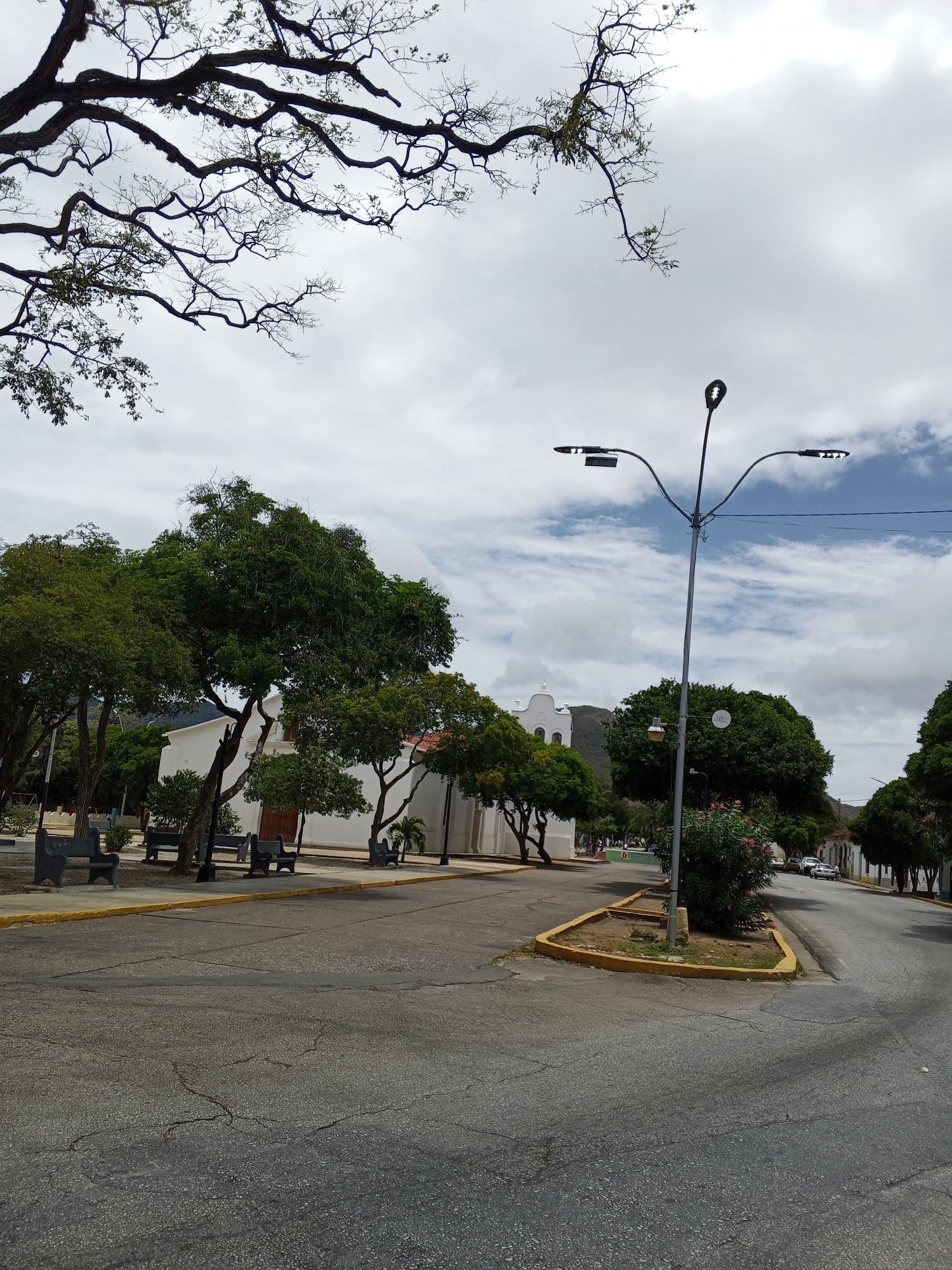 The image depicts a street corner in a town, possibly in a Latin American country given the architecture and vegetation. The street pavement is cracked and appears aged. On the left side, there's a park area with trees, benches, and a white building that may be a church or public building. A street lamp stands on the corner, and the sky is partially cloudy. The overall environment suggests a quiet, sunny day in a small town.