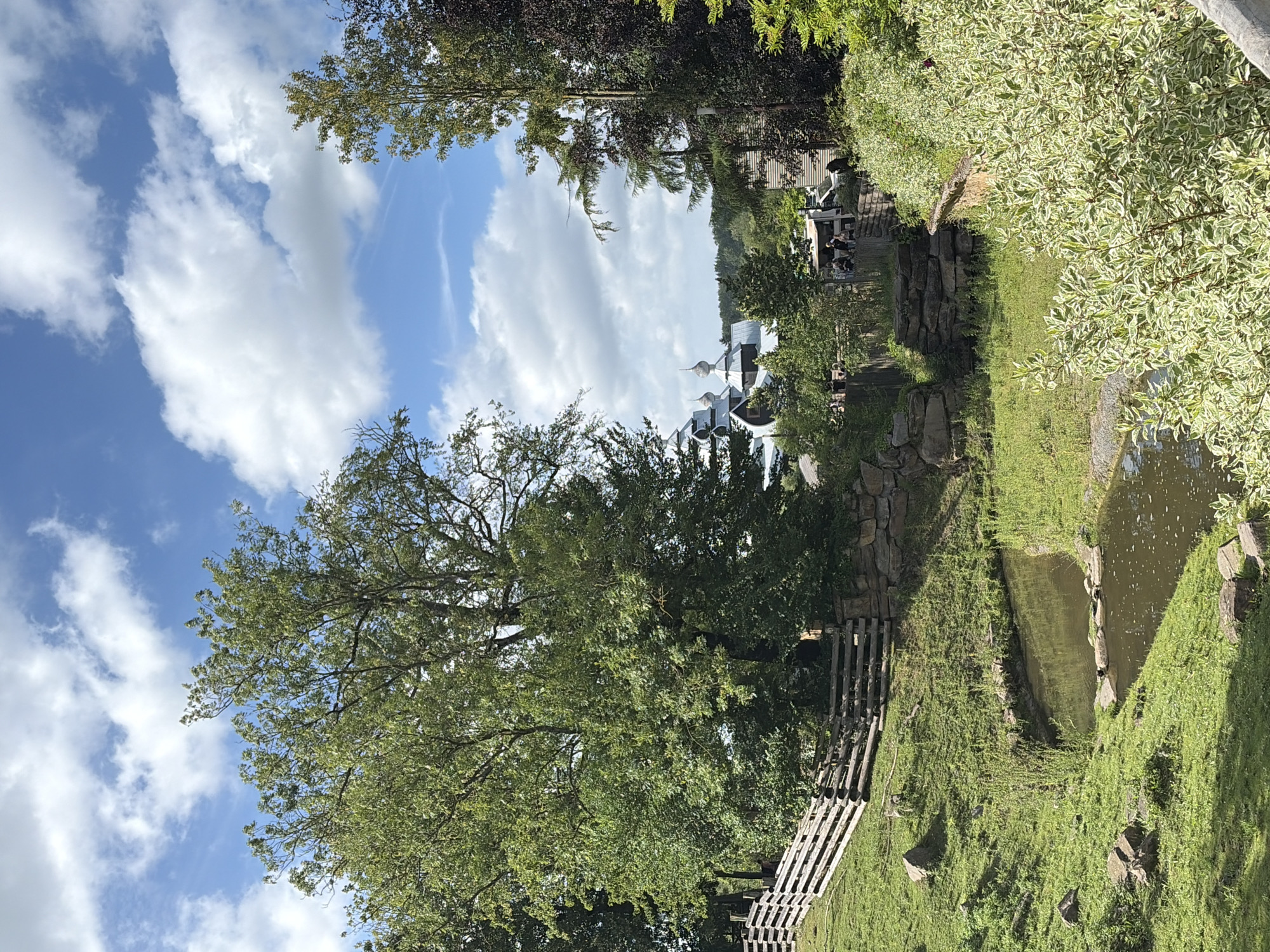 The image depicts a scenic view in Cambron-Casteau, Belgium, likely within a park or zoo setting. In the foreground, a small, winding stream flows through lush green grass. The banks of the stream are lined with various plants and rocks. A rustic wooden fence runs along the upper left of the scene, indicating a boundary or enclosure.
In the middle ground, several trees dominate the landscape, their dense green foliage creating a sense of depth. Behind the trees, glimpses of buildings can be seen, featuring distinctive domed roofs that suggest an architectural style, possibly of Eastern European or Russian influence. These structures appear to be part of an attraction or exhibit.
The background reveals a further expanse of trees and greenery, blending into a distant landscape. The sky above is a vibrant blue, adorned with fluffy white clouds, suggesting a pleasant, partly sunny day. A few people are visible in the distance, standing near the buildings, but their activities are indistinct. The overall impression is one of a tranquil outdoor environment with elements of both natural beauty and man-made structures.