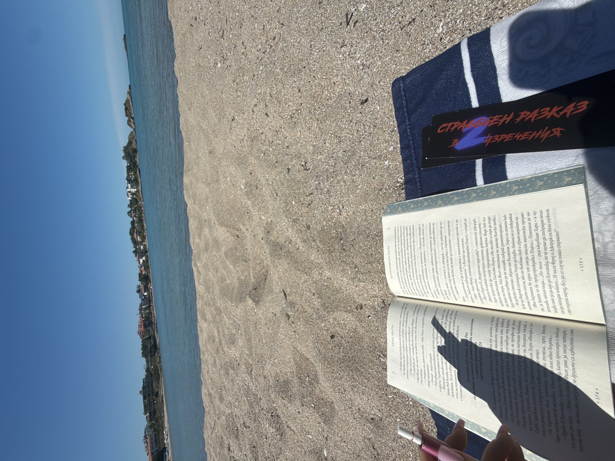 An open book rests on a blue and white striped beach towel on a sandy beach. The book is open to two pages of text, with the left page titled "ГЛАВА ЛЕГСТВА" and the right page titled "ХИТРОУМНАТА КАРТА". A pink object, possibly a vape or e-cigarette, is held in the foreground, casting a shadow over the left page of the book. A black bookmark with red and blue lettering is tucked into the right side of the book.
In the background, a calm, blue sea meets a light sandy beach. Beyond the beach, a small town with buildings of varying heights and colors is visible on a gentle incline leading to the sea. Several small rocky islands are dotted in the water. The sky is a clear, bright blue, suggesting a sunny day. The city of Tsarevo, Bulgaria, is the likely location, given the coastal scenery and architectural style.