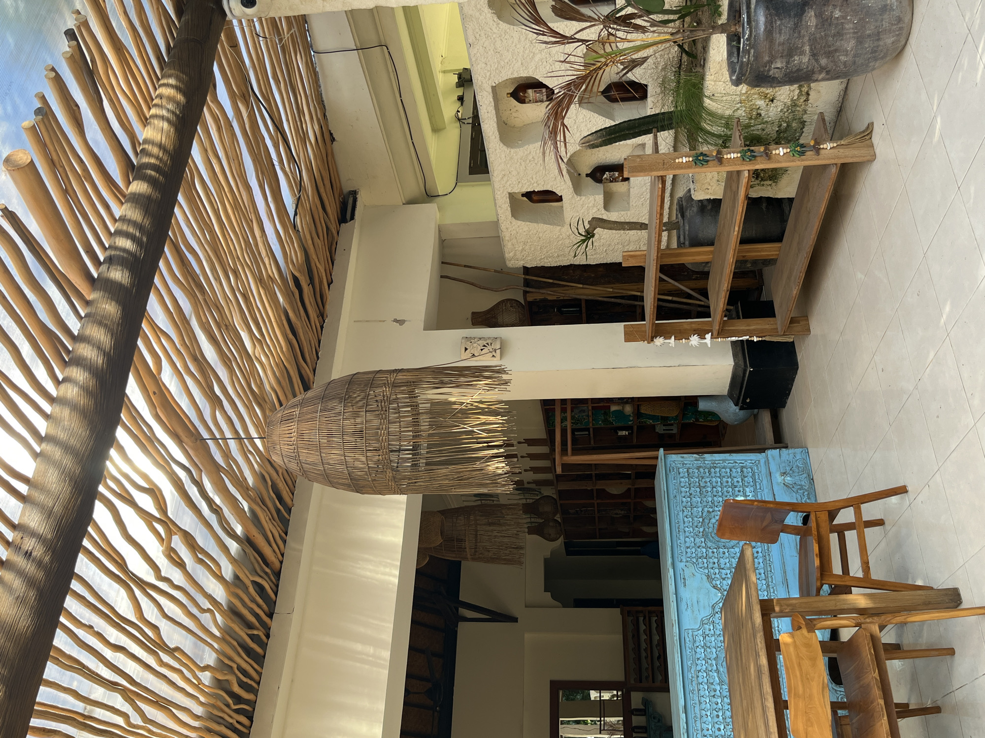 The image depicts an outdoor seating area at a venue in Kuta Selatan, Indonesia, likely a restaurant or cafe. The setting features a rustic, natural aesthetic with a slatted wooden roof that allows sunlight to filter through. Hanging from the ceiling are two large, woven pendant lights made of natural materials.
In the foreground, a wooden dining table is set with two wooden chairs. Behind the table is a distinctive bright blue, ornately carved sideboard or counter. To the right of the blue furniture is a rustic, multi-tiered wooden shelving unit displaying potted plants, including a tall cactus. The wall behind the shelves is textured and has built-in niches, some containing brown bottles.
Further into the background, through an open doorway, more shelving units are visible, stocked with various items. The floor is tiled with large, light-colored square tiles. The overall impression is one of a casual, bohemian-inspired outdoor space. There are no people visible in the image.