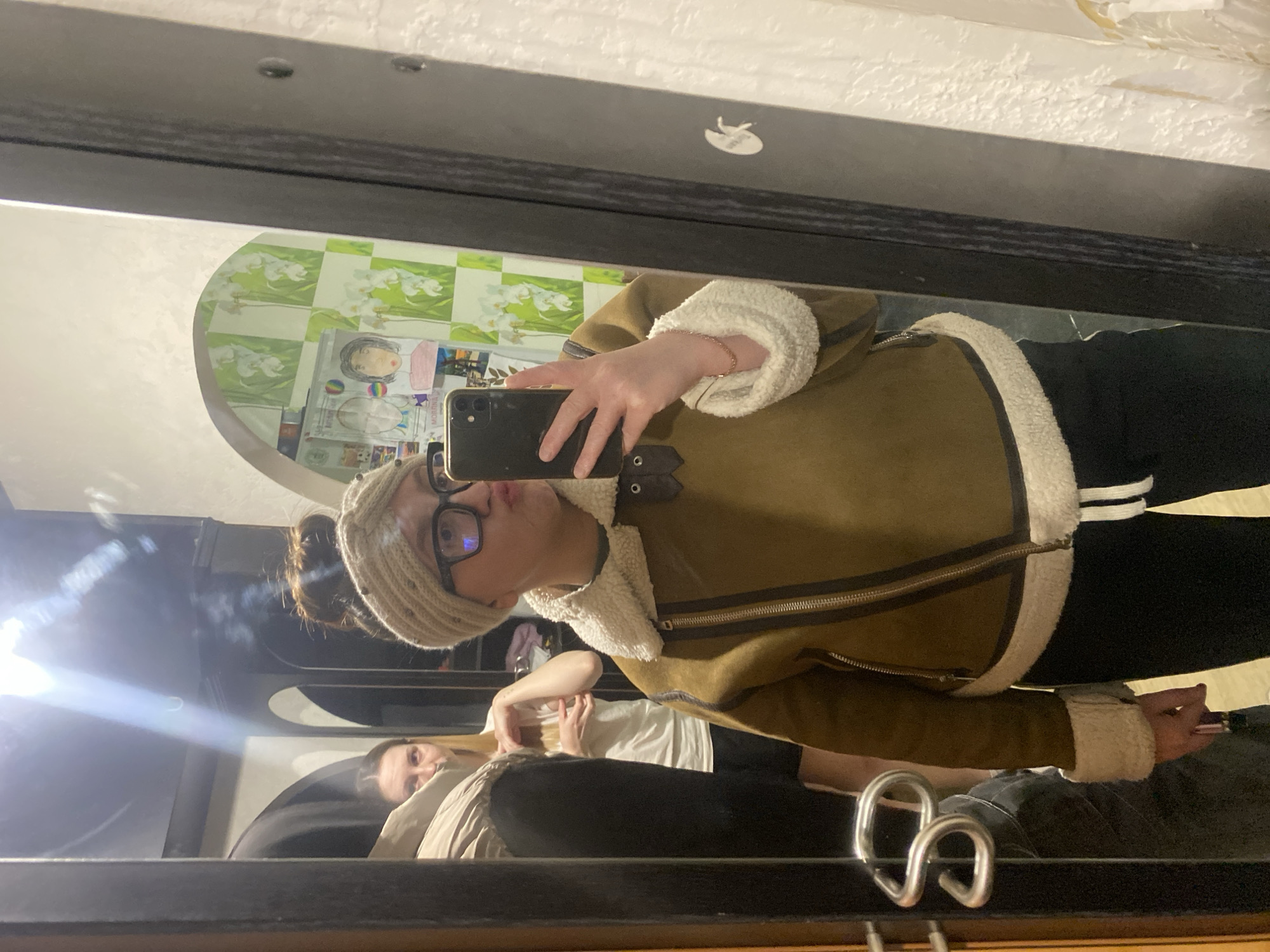 The image depicts an indoor scene in Chaikovsky, Russia, captured as a mirror selfie. A female individual, the primary subject, is reflected centrally, wearing a brown shearling-lined bomber jacket, black pants with white drawstrings, a beige knitted headwrap, and black-framed glasses. Her hair is pulled into a bun. She holds a black smartphone with a triple camera in her right hand, photographing her reflection while posing with pursed lips. A rose gold bracelet is visible on her left wrist. To her left in the reflection, a second female individual with long blonde hair, wearing a white t-shirt and dark shorts, stands with her arms bent, observing. The reflected background includes a wall with alternating green and white patterned wallpaper featuring white floral designs, and several taped drawings or posters, including two stylized faces. A dark-framed doorway or archway is visible on the far left, and a bright light source creates a prominent lens flare in the upper left quadrant of the mirror. A silver S-shaped door handle is partially reflected on the left edge.