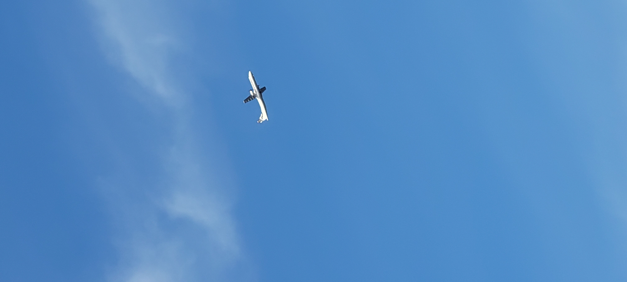 The image shows an airplane in flight against a clear blue sky with wispy clouds. The airplane, which appears to be a turboprop aircraft with two engines mounted on the wings, is white with dark markings on its tail and fuselage. It is captured in a side profile, ascending or cruising at a moderate altitude. The bright sunlight illuminates the aircraft from the side. There are no visible landmarks or features in the image that would allow for the specific identification of its location within Riga, Latvia. The environment is solely the sky and the airplane.