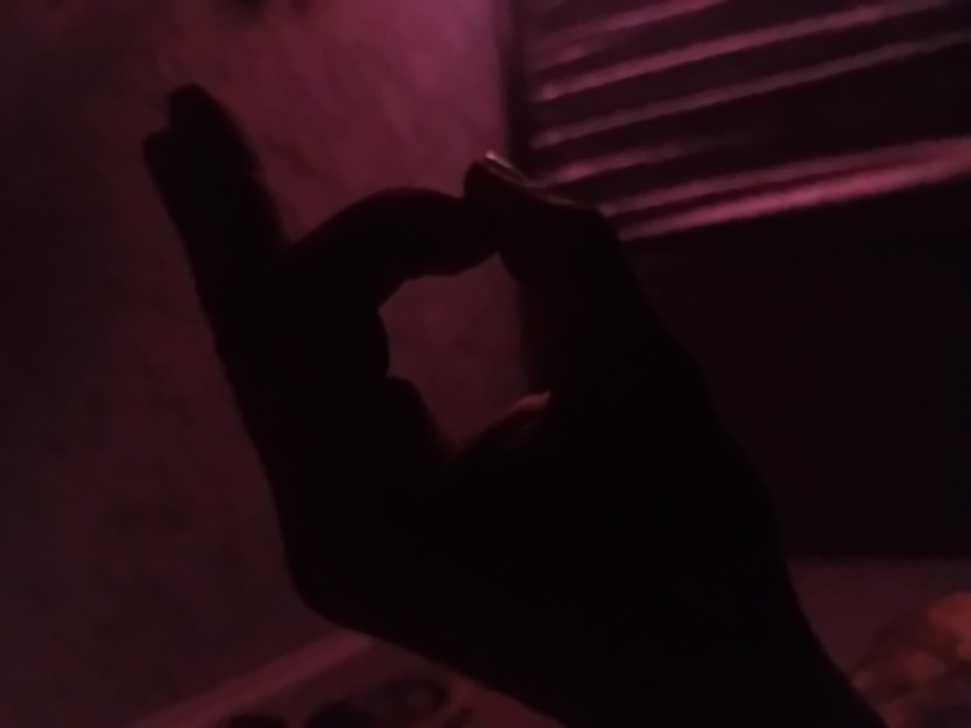 A silhouette of a hand forms an "O" shape against a dark, dimly lit background with pinkish hues. The hand is positioned in the foreground, with the thumb and index finger nearly touching, creating a circular opening. The background appears to be an interior space, possibly a room, with curtains visible on the right side. The lighting suggests an artificial light source, casting a subtle glow on the surroundings. The scene is devoid of any specific identifiable landmarks or context related to the city of Verkhniachka, Ukraine, and offers no discernible information about any individuals or actions taking place beyond the formation of the hand gesture.