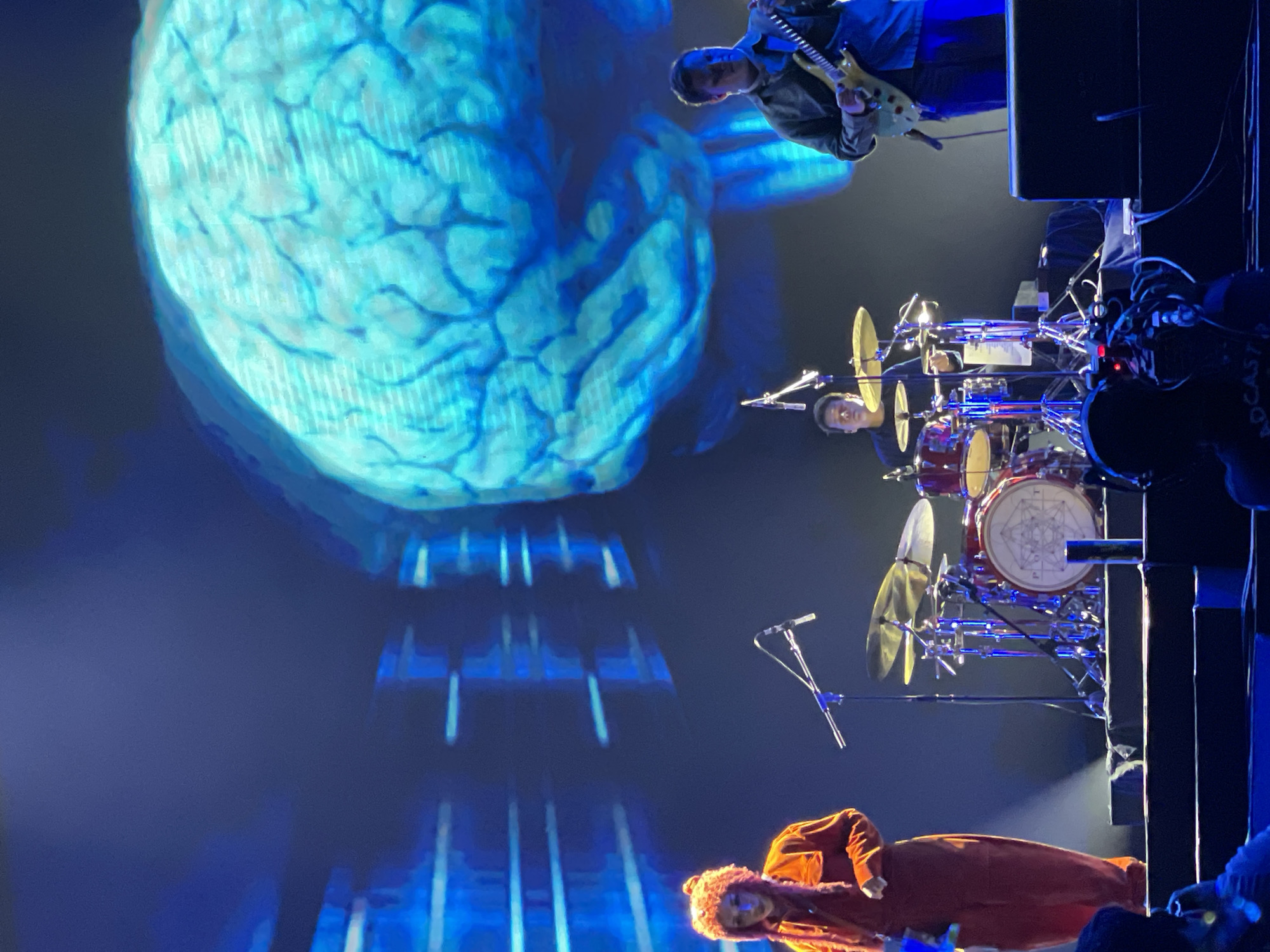 A band is performing on stage, bathed in blue light. A large projection of a brain is visible behind them. The band consists of a guitarist on the upper right, a drummer in the middle, and a vocalist on the bottom. The drummer is behind a drum set with cymbals and a red drum with a white design on it. The vocalist, wearing an orange jacket and hat, is lying on the stage. The location in Salembaran, Indonesia is likely a concert venue.
