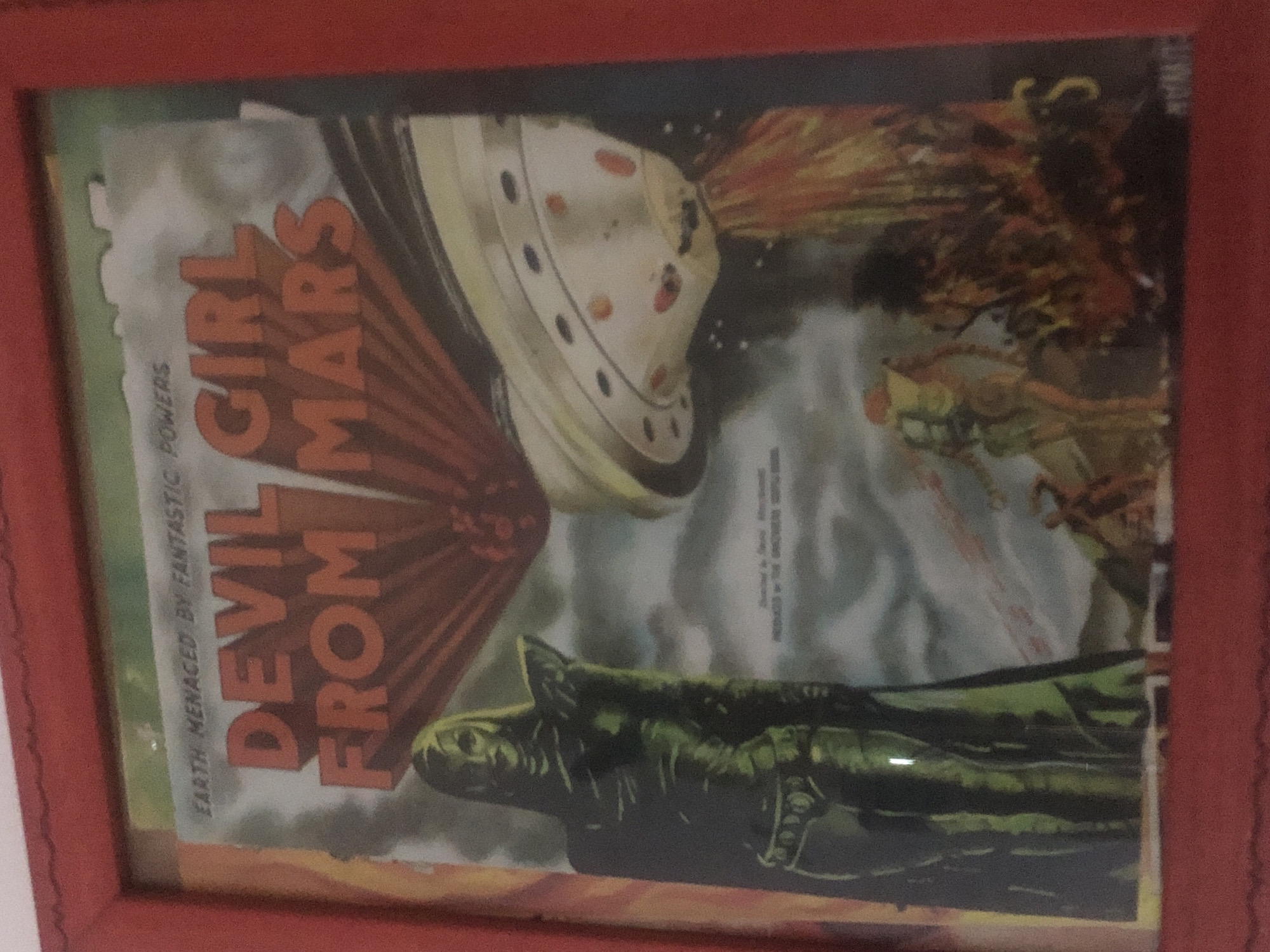 The image is a framed, close-up shot of a vintage comic book cover titled "DEVIL GIRL FROM MARS." The cover depicts a tall, green-skinned female figure in the foreground, dressed in a metallic-looking costume. Behind her, a flying saucer hovers in a cloudy sky, emitting beams of light. The background shows a desolate, alien landscape with two smaller figures in the distance, one appearing to be fleeing. The comic cover is framed in a red border. The image quality is slightly blurred, suggesting it may be a photograph of the framed item. The specific location within a city is not discernible from the image.