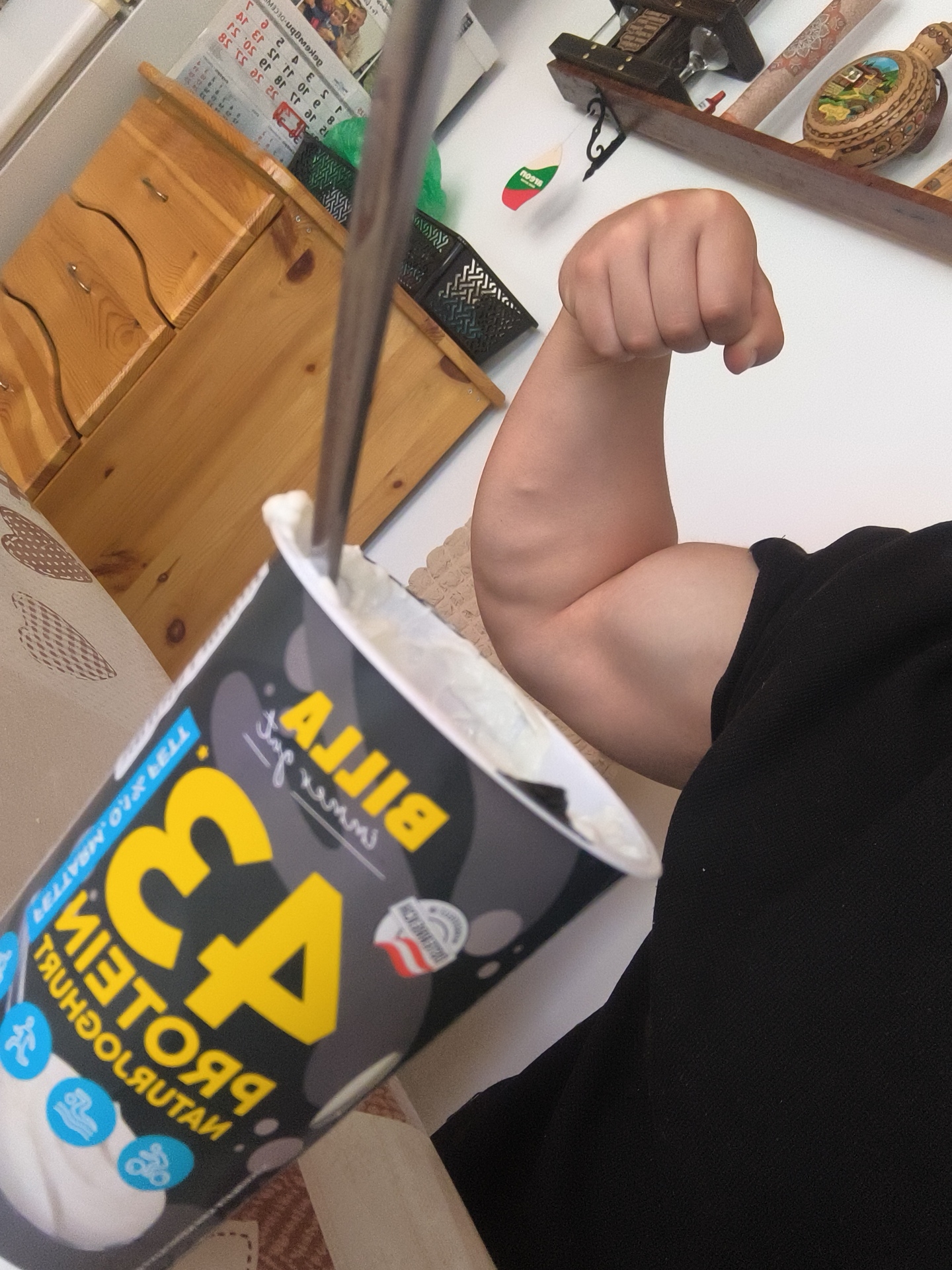 The photograph shows a close-up shot of a person's flexed bicep, presumably showing off their strength. In the foreground is a container of "Billa 43 Protein Natur" yogurt with a spoon inside. The brand name "BILLA" is visible. A wooden shelf adorned with decorative items and a calendar can be seen in the background. The person is wearing a black garment. The yogurt brand indicates that the photograph was likely taken in Bulgaria, as "BILLA" is a supermarket chain present in the country. The location within Bulgaria is not possible to determine to Malka Smolnitsa, Bulgaria.