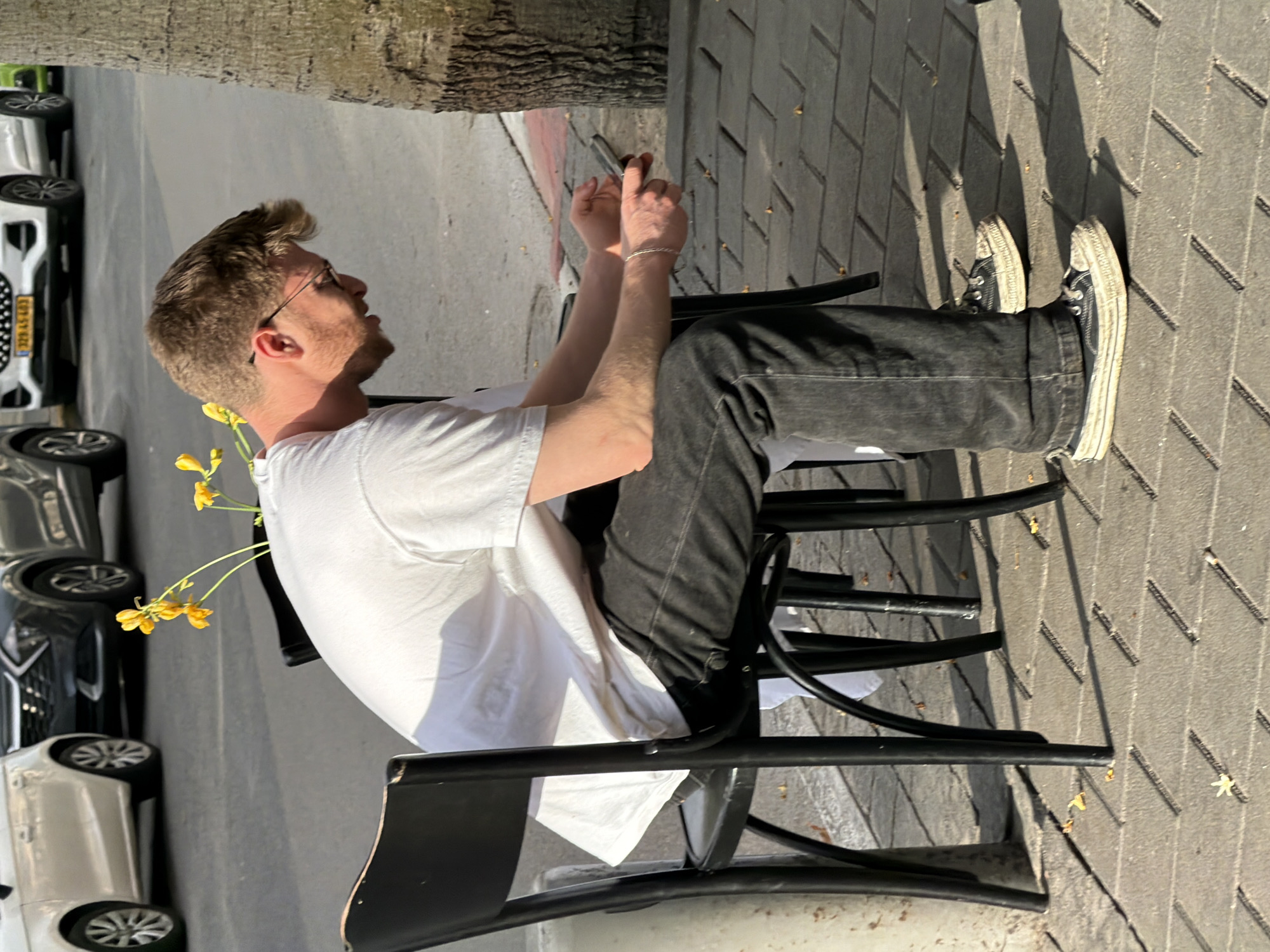 The image presents a daytime scene on an urban sidewalk in Tel-Aviv, Israel. A male individual, appearing to be in his 20s or 30s, is seated on a black wooden chair facing left, his body angled towards the viewer. He has light brown hair, a short beard, and wears wire-frame glasses. His attire consists of a white short-sleeved t-shirt, dark (likely black or charcoal) jeans, and worn black canvas shoes with white soles. A silver-colored braided bracelet is visible on his left wrist. He is actively engaged with a dark-colored smartphone, holding it horizontally with both hands and looking down at its screen.
Behind the man, a small cluster of yellow flowers with green stems is visible, positioned as if on a table surface. The sidewalk is paved with rectangular grey interlocking tiles, with some small, light-colored debris scattered on the ground. To the immediate right, a tree trunk with textured bark rises from the sidewalk. A red-painted curb delineates the sidewalk from an asphalt street in the background. Several vehicles are present on the street: a prominent white SUV with Israeli license plate "329-LS-483" is parked behind the man, flanked by a dark grey SUV partially visible to its left and a white sedan on the far left. A glimpse of a green vehicle is visible on the far right. The scene is illuminated by strong sunlight, casting defined shadows.