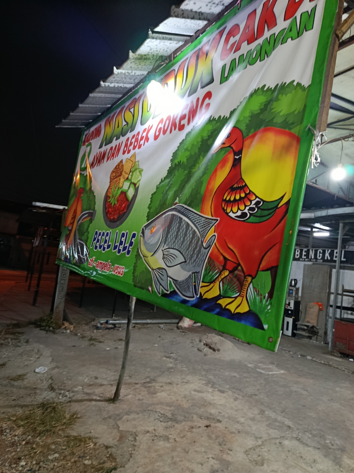 This is a nighttime shot of a banner advertising a food stall in Depok, Indonesia. The banner is predominantly green and features cartoonish illustrations of food items and animals.
The main subjects on the banner are a red and yellow duck, a grey fish, a plate of food that appears to be rice with chicken, vegetables, and sauce, and an illustration of a mango. There is also a smaller illustration of a chicken leg and some text below it. The background of the banner depicts a green, grassy landscape with a blue sky and some yellow elements, possibly representing more fruit or sunlight.
The banner is supported by two wooden poles and is partially illuminated by artificial lighting. The lighting suggests it is nighttime. In the background, beyond the banner, there are some buildings and structures, including a sign that reads "BENGKEL" which means "workshop" in Indonesian, and a phone number. The ground in front of the banner is paved, with some patchy grass and debris.
Visible text on the banner includes:
- "NASI AYAM DAN BEBEK GORENG" (Rice, Fried Chicken and Duck)
- "LAMONGAN" (Likely referring to the origin or style of the food)
- "PECEL LELE" (Fried Catfish)
- "ati-ampela-usus" (Liver, Gizzard, Intestine - possibly side dishes)
The overall impression is of a typical roadside food vendor in Indonesia, operating in the evening or at night. The banner is colorful and designed to attract customers.