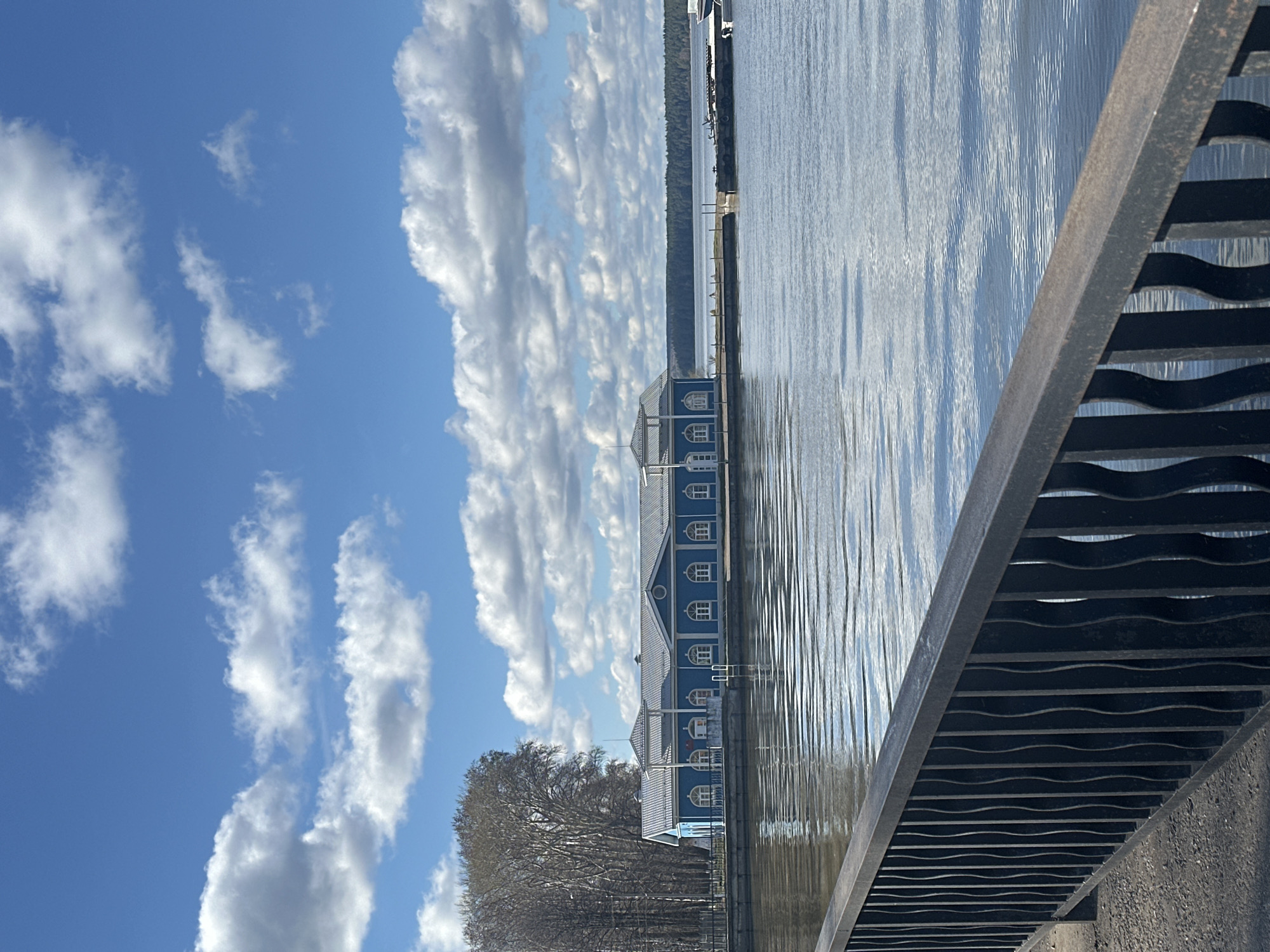 The image shows a body of water with a building alongside it, likely located in Votkinsk, Russia. The building is blue with white windows and multiple stories, suggesting it might be a boathouse or another type of facility related to water activities. The presence of a bridge with a metal railing is visible in the foreground, partially obscuring the view. The sky is bright blue with scattered white clouds, suggesting a sunny day. The water is reflecting the sunlight, creating shimmering patterns on the surface. Some trees are visible on the shore near the building.