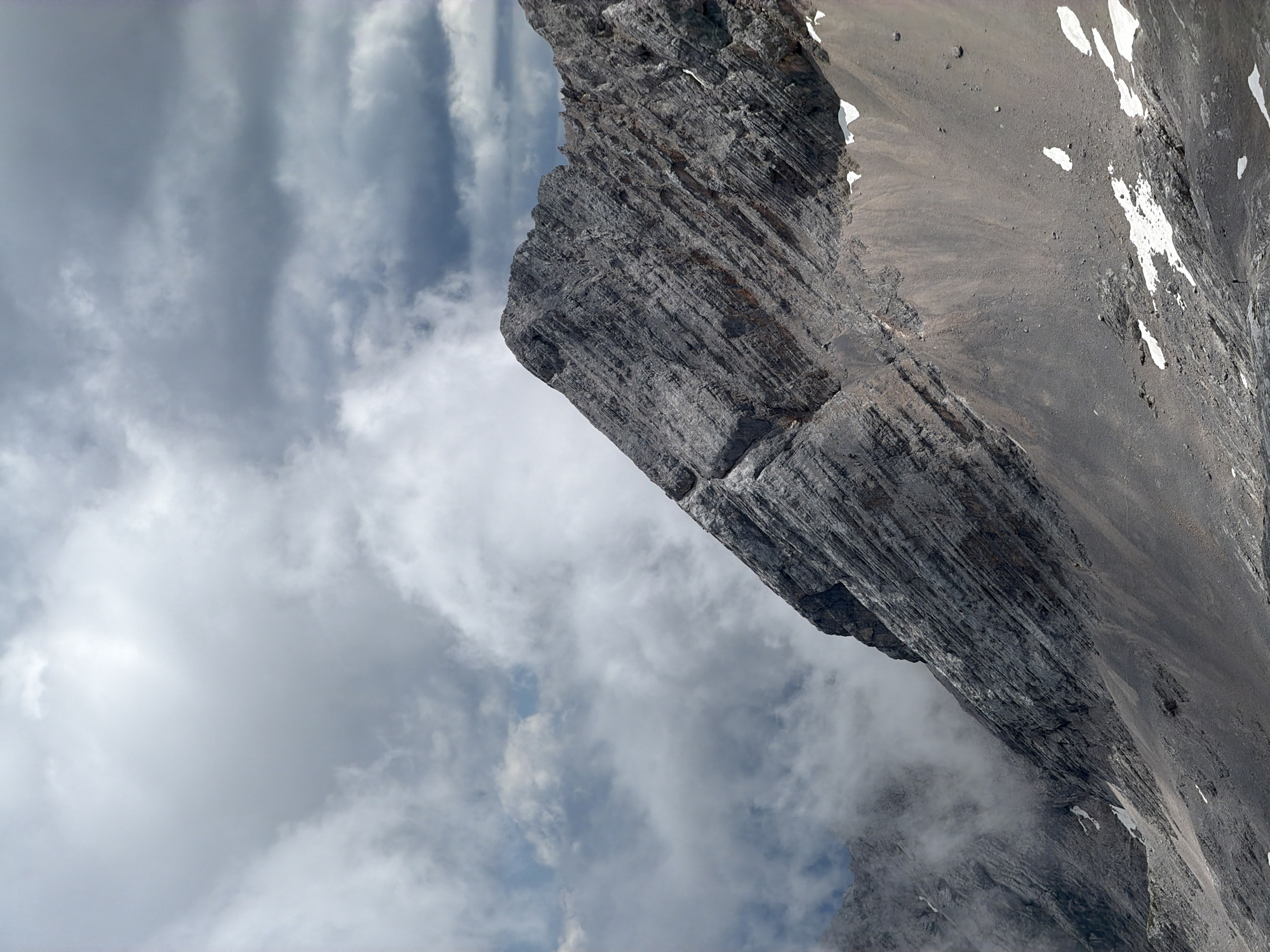 The image shows a dramatic mountain landscape with rugged, gray rock formations dominating the foreground. The mountain slopes are steep and textured with layers of rock, indicative of geological stratification. Patches of snow are visible on the lower slopes, contrasting with the rocky terrain. The sky above is filled with heavy, dark clouds, suggesting an overcast or stormy weather condition. The overall scene conveys a sense of a high-altitude, natural environment. This scene is likely a view of the Zugspitze massif, Germany's highest peak, located in the Wetterstein Alps near Garmisch-Partenkirchen. The image captures the imposing nature of the mountain range under a dramatic sky.