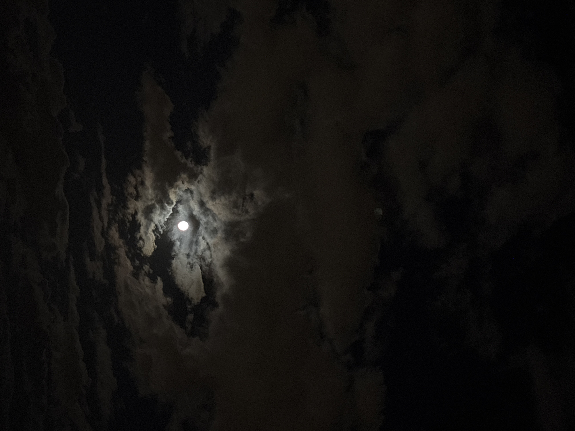 The image depicts a night sky with a bright full moon visible through breaks in a thick cloud cover. The clouds are dark and textured, creating a dramatic and somewhat ominous atmosphere. No people, buildings, or other city-specific features are discernible. The lack of distinguishable foreground elements prevents confirmation of the location in Minsk, Belarus.