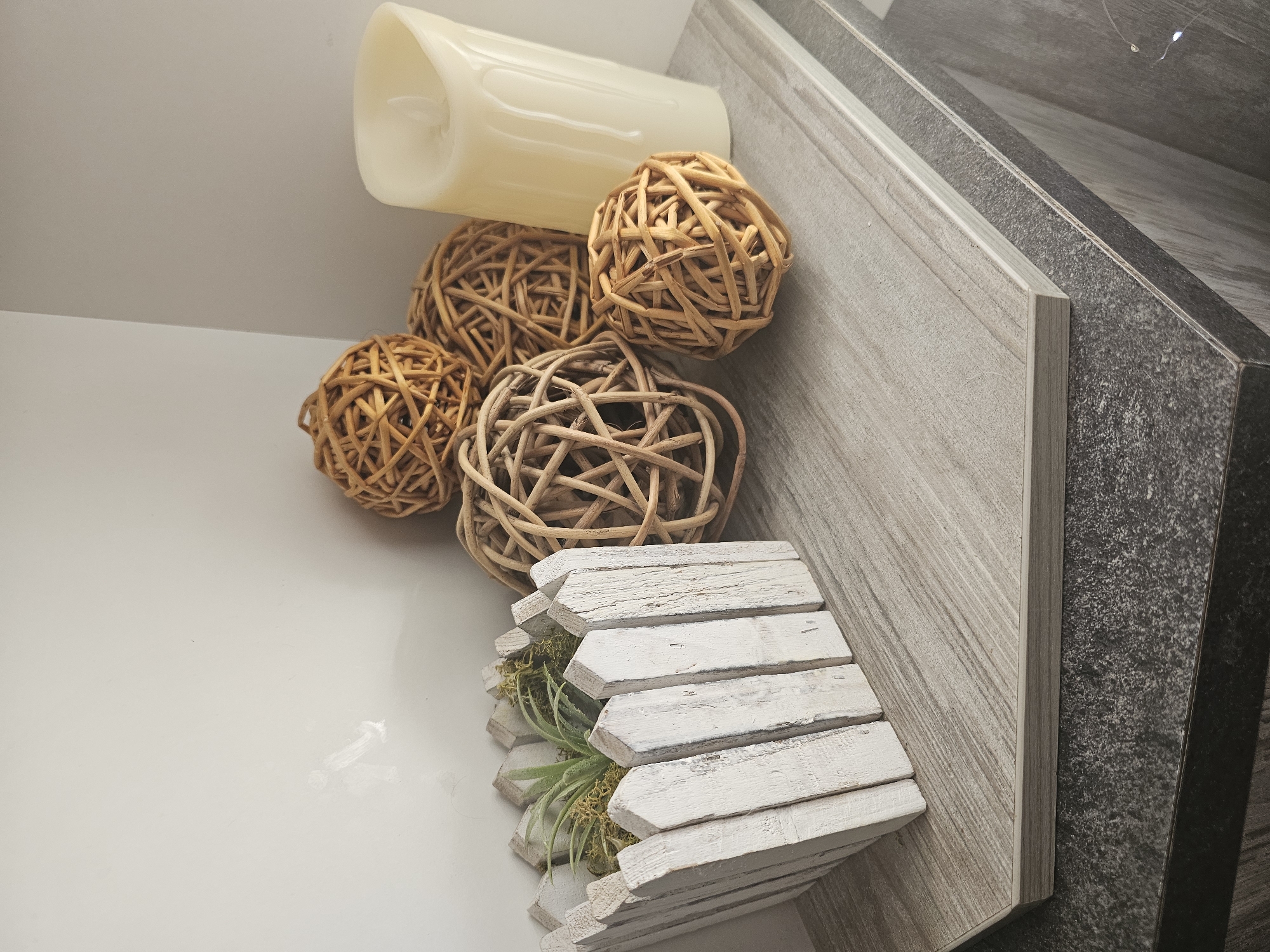 The image displays a collection of decorative items arranged on a smooth, white surface next to a tiled background. The primary subjects are several spherical, woven decorative balls made of natural material, a tall, cream-colored faux candle, and a small, white wooden planter housing a green succulent plant and moss. The planter has a unique hexagonal shape with pointed, overlapping wooden slats.
The objects are artfully arranged, suggesting a decorative vignette. The faux candle is positioned at the top left, partially obscuring some of the woven balls. The balls are clustered together, with varying sizes and textures. The wooden planter with the succulent is placed at the bottom right, showcasing its rustic design.
The background features a tiled surface with a light, wood-grain pattern on a diagonal, next to a darker, speckled tile. This suggests an indoor setting, possibly a shelf, tabletop, or a part of home decor. There is no visible activity or event taking place, as the image focuses on a still life arrangement.
Notable details include the texture of the woven balls, the matte finish of the faux candle, and the distressed, painted look of the wooden planter, which gives it a weathered appearance. The lighting appears to be soft and even, casting subtle shadows, indicating it might be daytime or a well-lit indoor space. There are no discernible location cues beyond the implied indoor setting, and no text is visible in the image. The overall mood is one of calm and aesthetic arrangement.