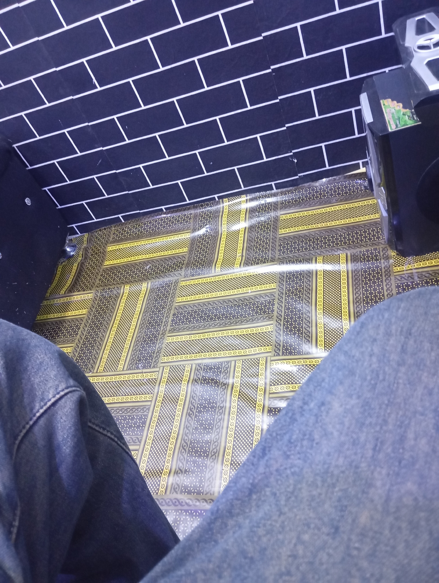 The image displays a floor covered with a patterned mat, featuring yellow and grey geometric designs. A black brick-patterned wall is visible in the upper left portion of the frame, with a speaker or electronic device on the right side. The foreground shows a person's legs clad in blue denim jeans, indicating they are seated or lying down. The scene appears to be indoors, with no clear indication of a specific city or location.