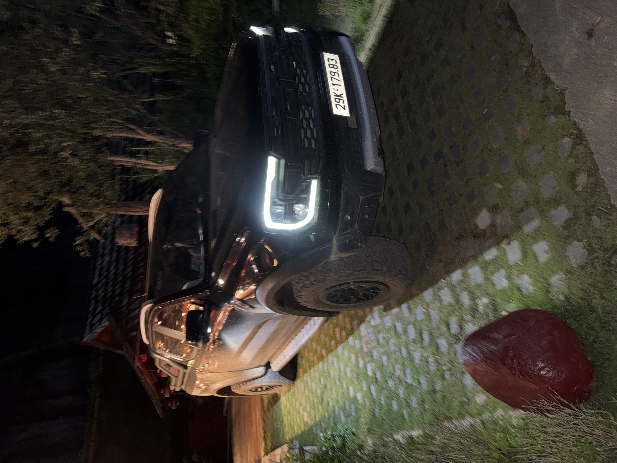 A black Ford Ranger pickup truck is parked at night in front of a building with a distinctive tiered roof, possibly in Moc Chau, Vietnam. The truck's headlights are illuminated, casting a bright glow on the front grille and the textured, square-patterned pavement. To the left of the truck, a red, roughly ovoid object sits on a patch of grass interspersed with paving stones. The background features dark trees and foliage, suggesting a natural or semi-rural setting. The overall lighting is dim, typical of nighttime conditions, with artificial light from the truck and the building providing illumination. There are no people visible in the image.