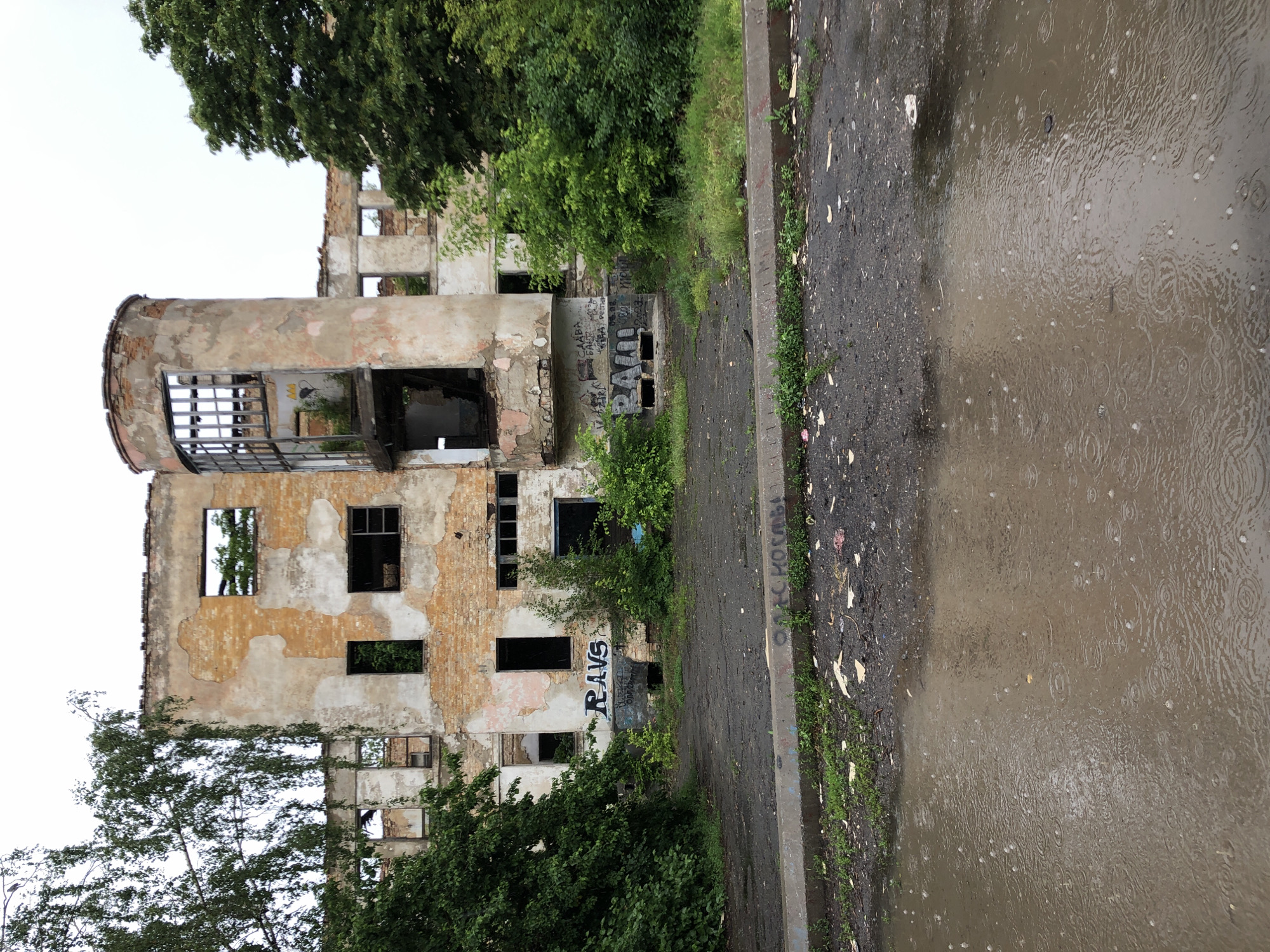 An abandoned, two-story building stands in disrepair, its brick facade crumbling and windows broken. The building has a circular tower on its right side. Graffiti covers some of the walls. Overgrown trees and bushes surround the structure. In the foreground, a muddy puddle reflects the overcast sky, with raindrops creating ripples on the surface. The ground is littered with debris.