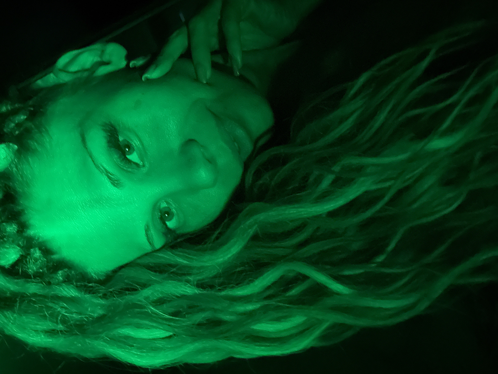 A close-up, head-and-shoulders shot captures a young woman with dreadlocks illuminated by vibrant green lighting. Her gaze is directed towards the camera, with her right hand resting on her cheek and chin. The woman appears to be in a dimly lit indoor setting, possibly a room, with the background fading into darkness. The image does not contain any specific identifiable landmarks or clues that would place it in Rusanovo, Russia.